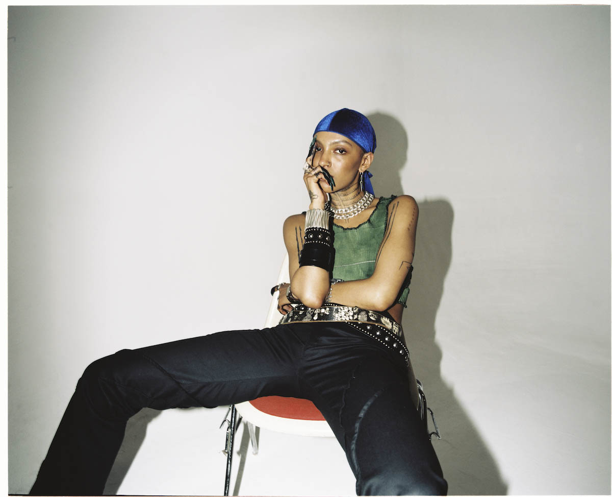 A female person sits broad-legged on a chair, looking directly into the camera. She wears a blue headscarf tied at the nape of her neck. She puts her right hand to her face, her little and ring fingers are bent under her nose, and she leans her index and middle fingers against her temples. The person wears a green sleeveless and cropped top, black pants, two wide belts, bracelets covering her right forearm almost to the elbow, two necklaces with large links on her neck. Tattoos are visible on the arms and neck.