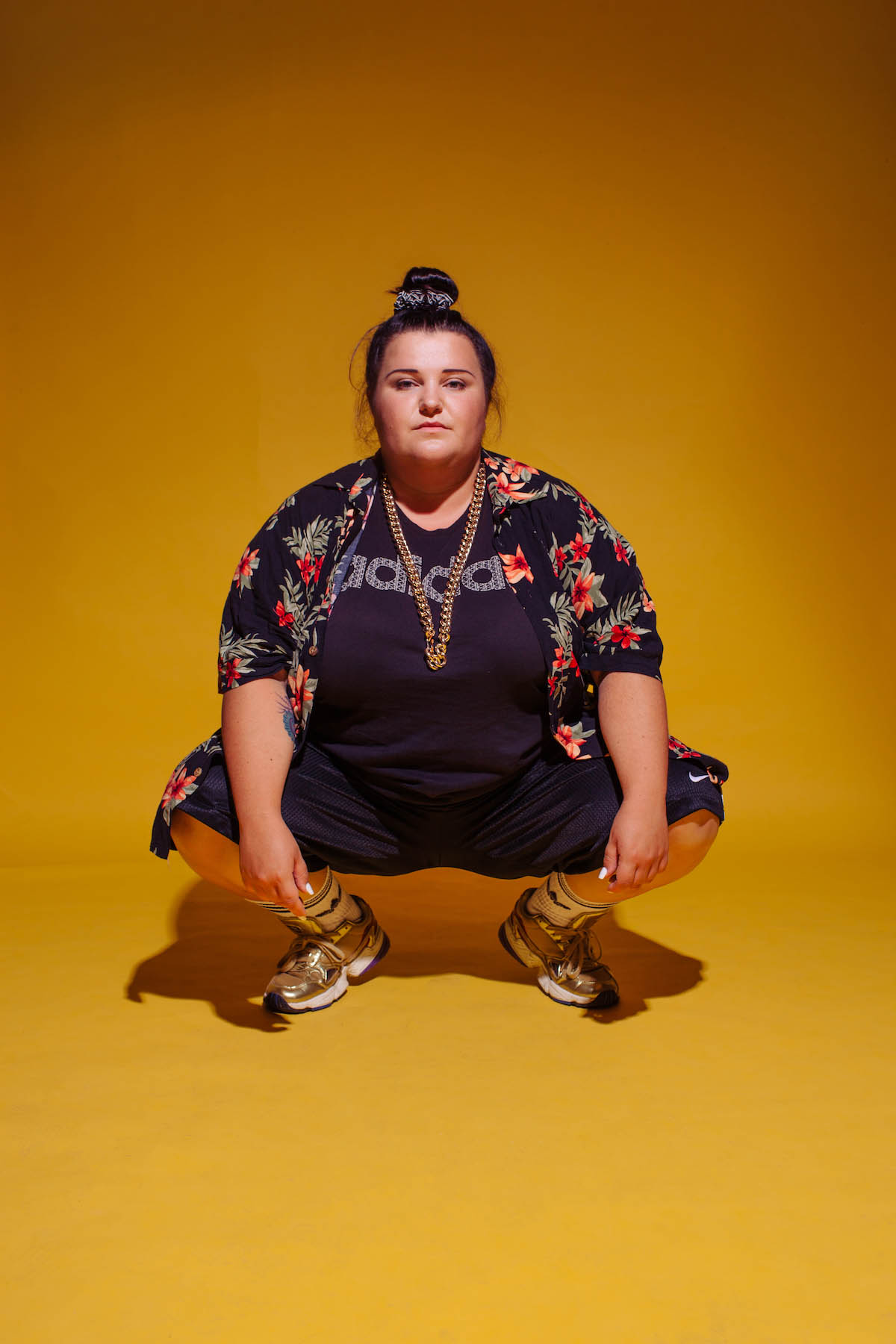 White, female person can be seen from the front in a squatting position. She is wearing a black T-shirt with Adidas written on it, a heavy gold chain that reaches her belly. Over the T-shirt she is wearing a black shirt with small red flowers on it, black knee-length trousers, white socks and gold-coloured trainers with white soles. Her hair is tied into a bun at the top of her head.