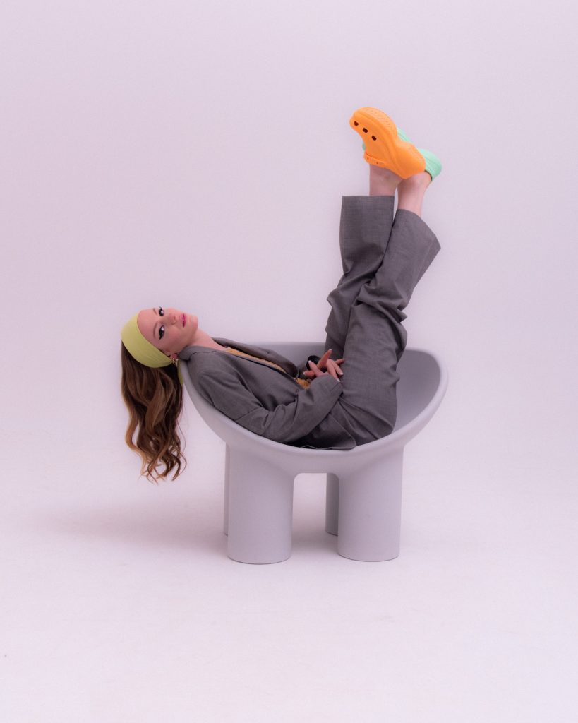 Young white woman lies sideways in a gray chair with semicircular large seat and thick chair legs. Her neck is resting on the left armrest, her long hair is hanging down beside the chair, her legs are both stretched straight up and crossed. She is wearing a wide yellow hair band, a gray suit, and two different colored Crocs shoes (orange and mint green). She is looking at the camera.