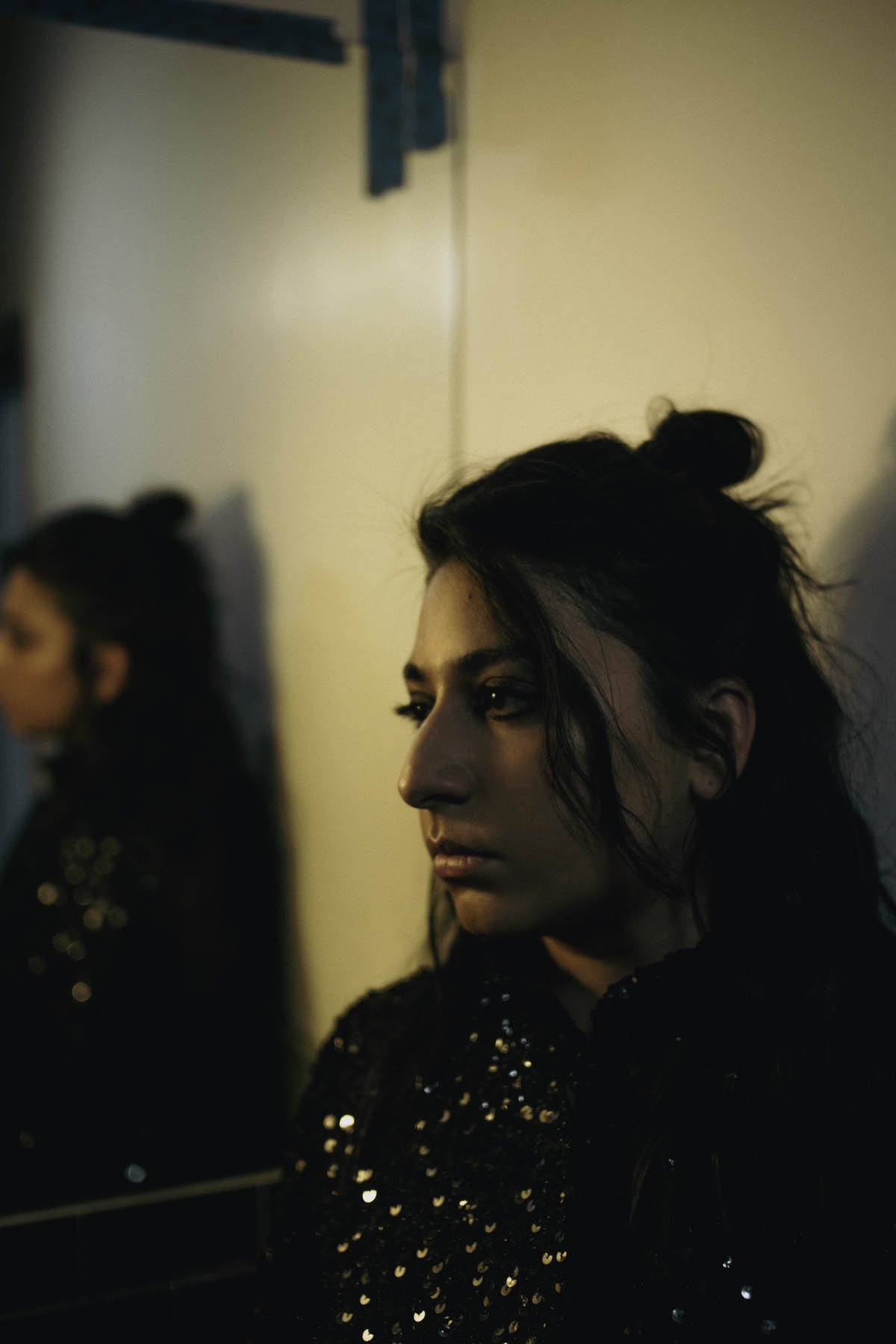 A female person can be seen in a semi-close-up. She wears her black hair tied into a small chignon on the top of her head, long strands fall into her face. Arooj Aftab wears a black top with sequins and collar and looks out of the picture to the left. Behind her hangs a mirror in which her image can be seen out of focus.