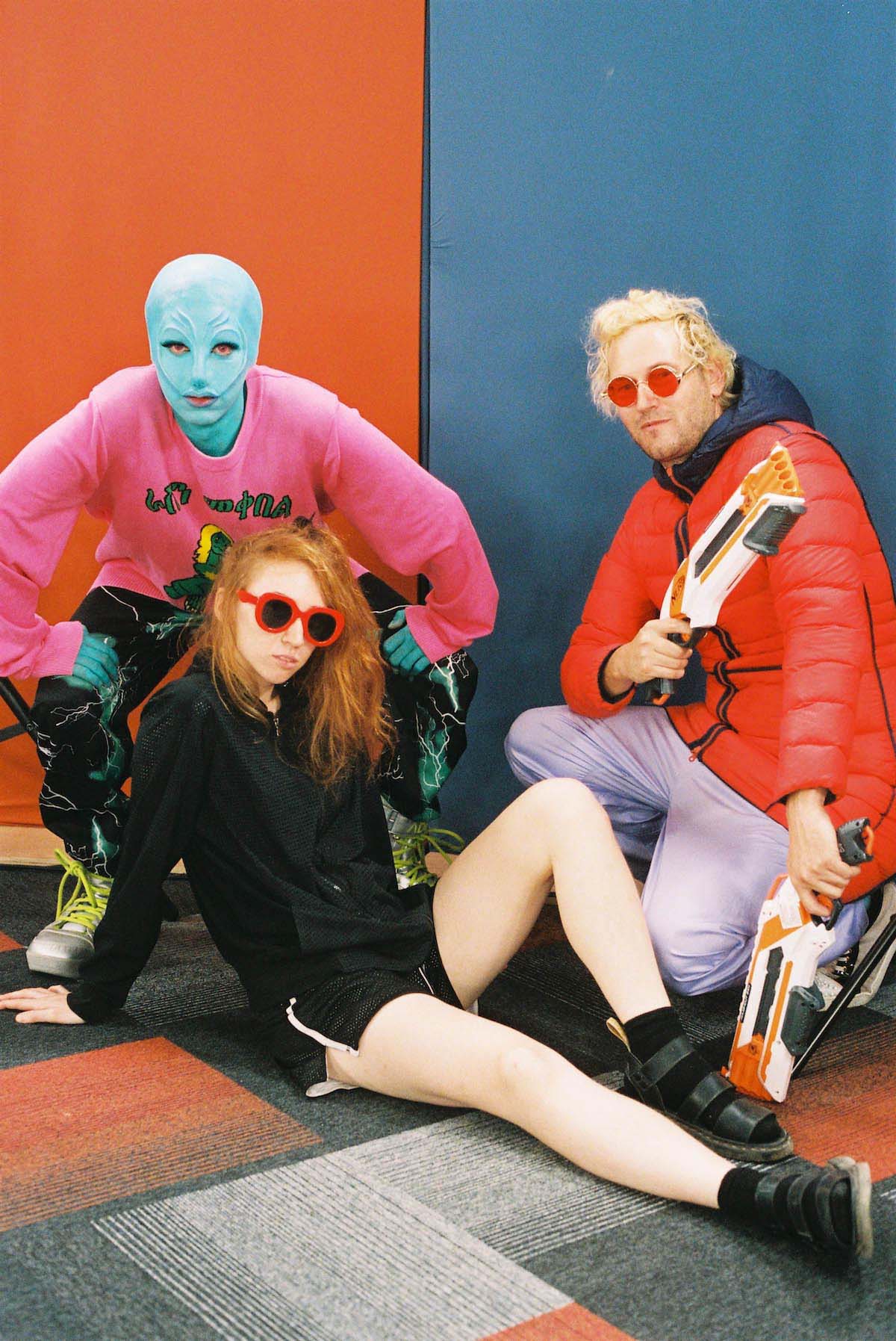 Three people, two squatting, one sitting on the floor. One person wears blue, alien make-up. The other two - a man and a woman - wear sunglasses. The woman has long red hair, the man short blond hair. He has two so-called Nerf Guns in his hands.