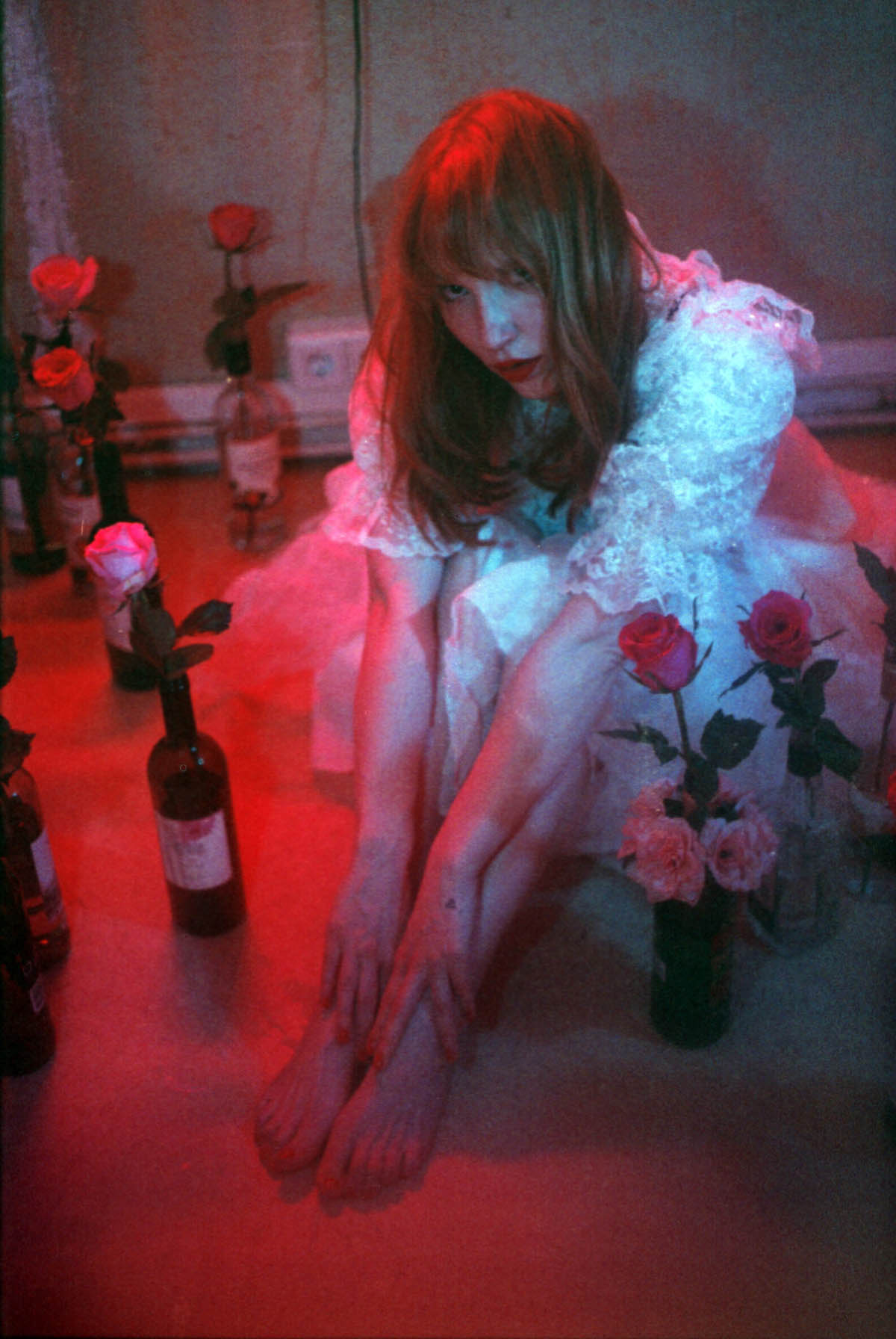 White woman with red hair and a white frilly dress sits bent forward on the floor. Her legs are bent, she touches her bare feet with her hands. She looks into the camera. Around her are single roses in several wine bottles.
