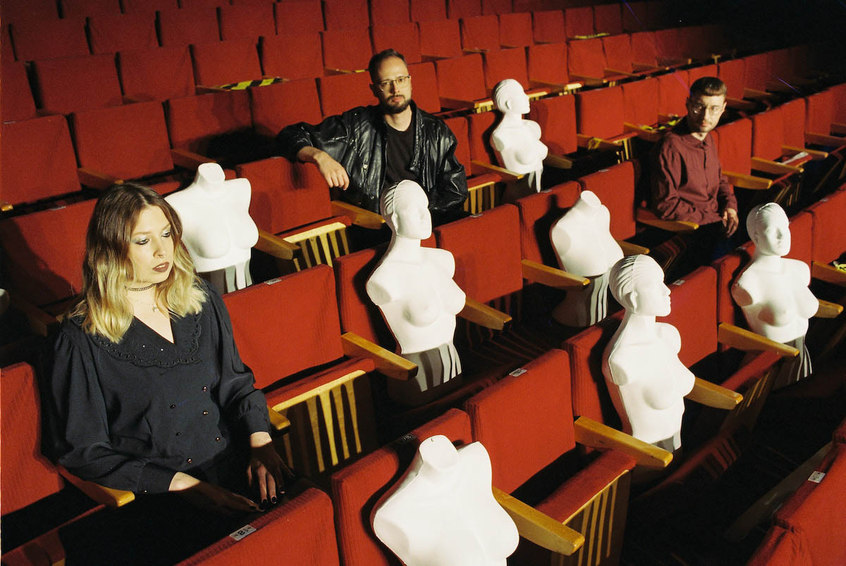 A woman and two men sit in an auditorium with folding seats with red cushions. Between and in front of them are several white busts of mannequin-like female torsos draped, some with heads, some without. The woman has blond hair slightly longer than shoulder length and stares forward toward the stage. One of the men sits in the same row as her further to the right with two busts between them. He is looking at the woman. In the row behind, a man in a black leather jacket sits between two busts. He looks at the camera.