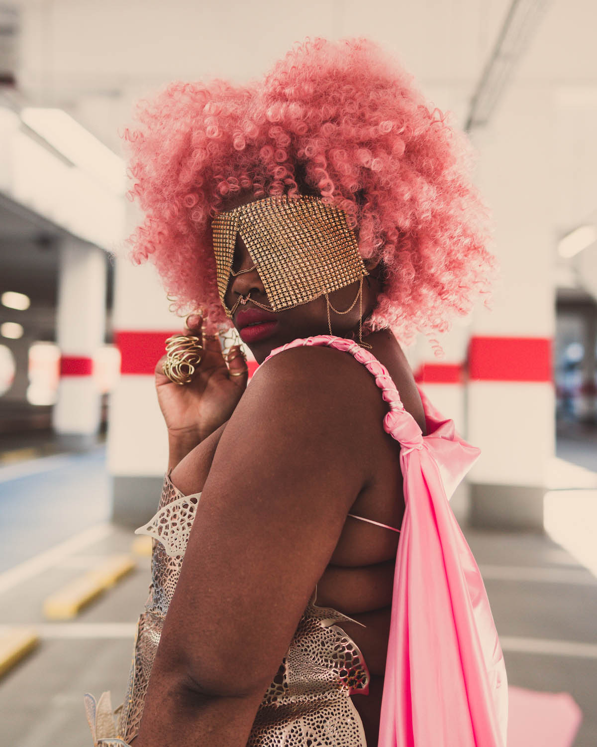 Female Black person is seen from the side down to the hip looking to the left. Donia Touglo wears curly pink hair. Her outfit consists of an equally pink cloak hanging down her back and an otherwise backless and sleeveless gold-coloured top that looks like the plant structure of a leaf with many holes of different sizes running through it. She shows a lot of skin. Donia Touglo holds her right hand at chin level. She wears gold rings that look like a spiral and stretches over several fingers. Her left and right halves of her face are covered except for her nose by two large square gold accessories that resemble blinders. Her golden nose ring is connected to these accessories by thin gold chains. She is standing in a multi-storey car park, which can be seen blurred in the background.