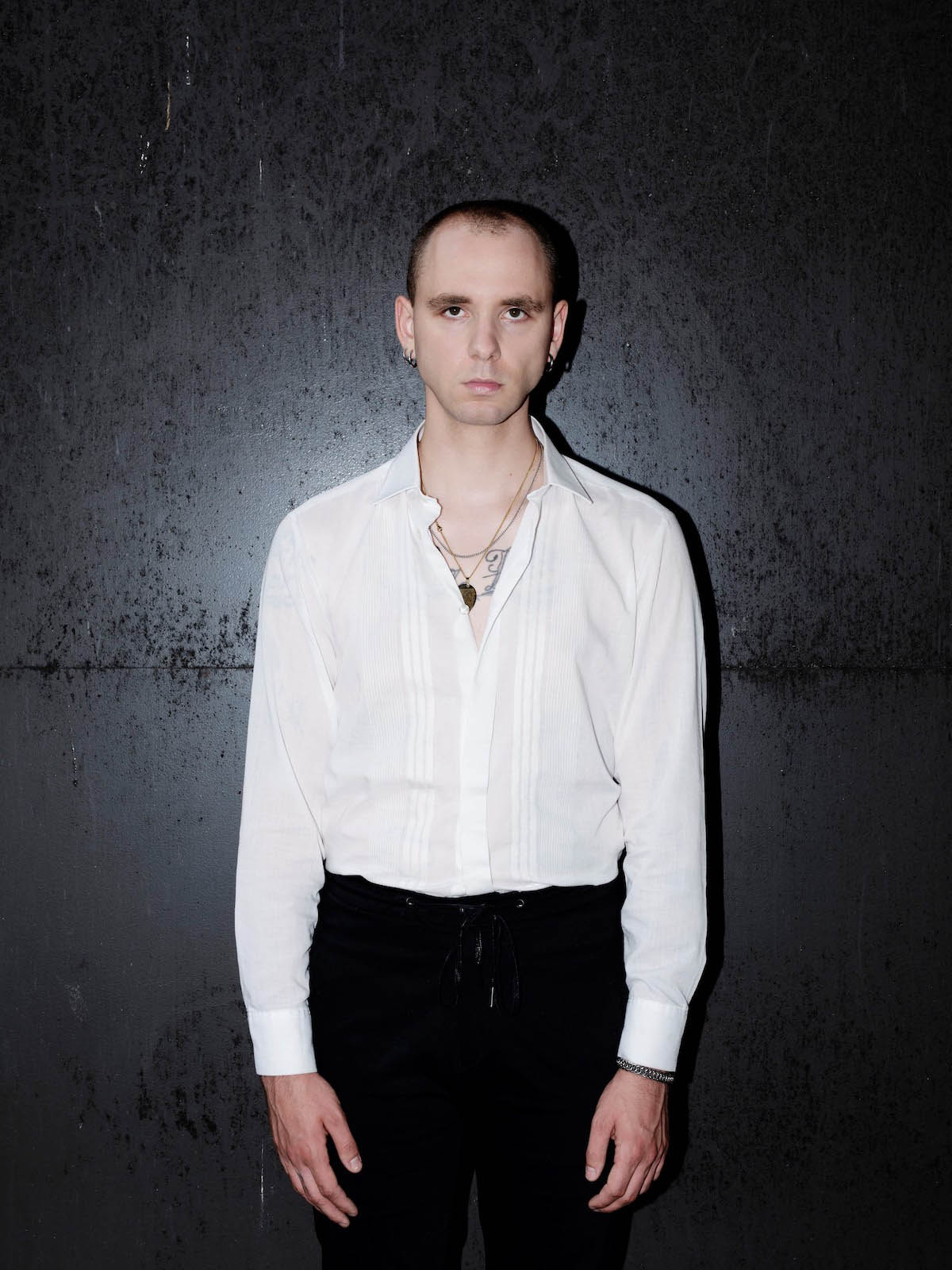 Male white person standing up visible down to the knees, looks directly into the camera, has very short hair, wears a white shirt and black trousers. The top buttons of the shirt are unbuttoned, so you can see a necklace and tattoos. He is wearing earrings in both ears.