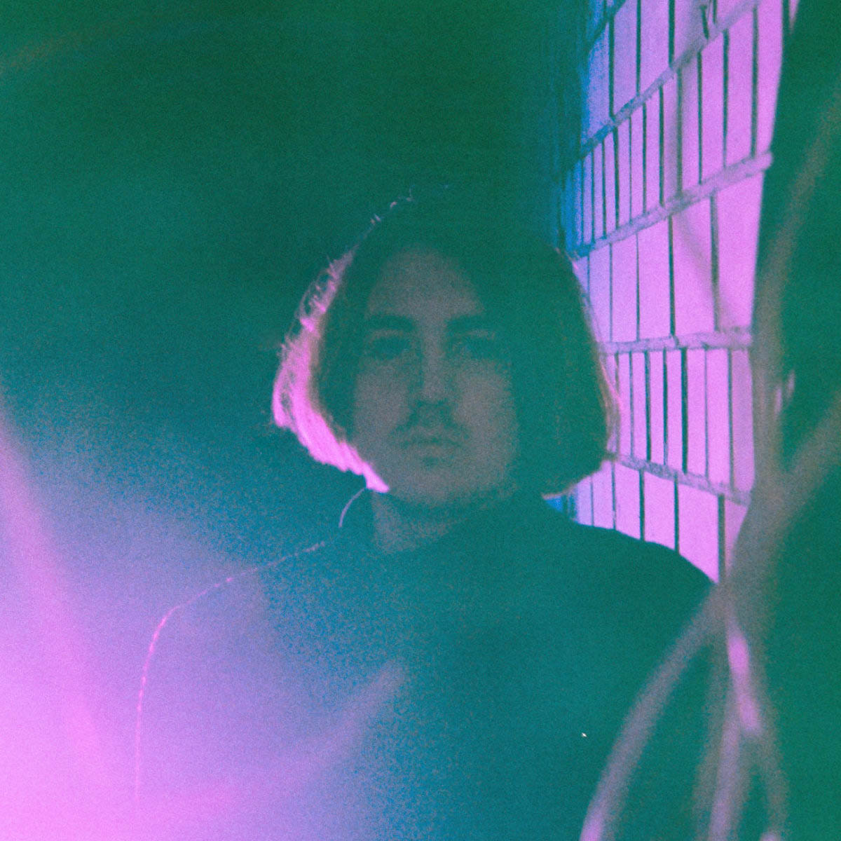 Blurred photo of a young man with chin-length hair and a moustache. Edward Hunt wears a jumper with a high collar and looks into the camera. He can be seen up to his chest. There is a stone wall behind him on the right. Part of his hair and the wall are bathed in purple light. A purple glow also emanates from the bottom left corner of the picture. The rest of the picture is black and rather gloomy.