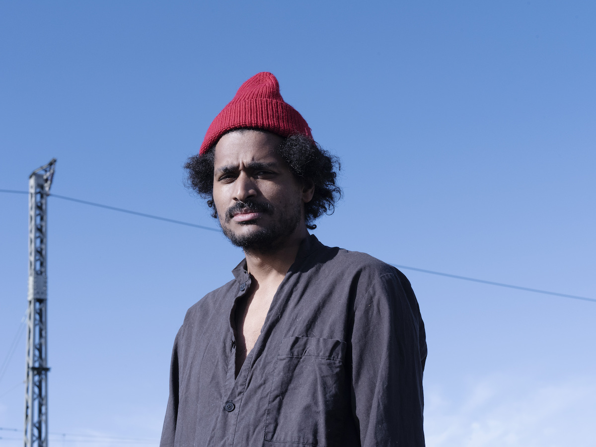 Person of Colour with black hair, black upper lip and chin beard, grey shirt and wine-red beanie cap. Fehler Kuti looks into the camera and squints his eyes. Blue sky can be seen in the background.