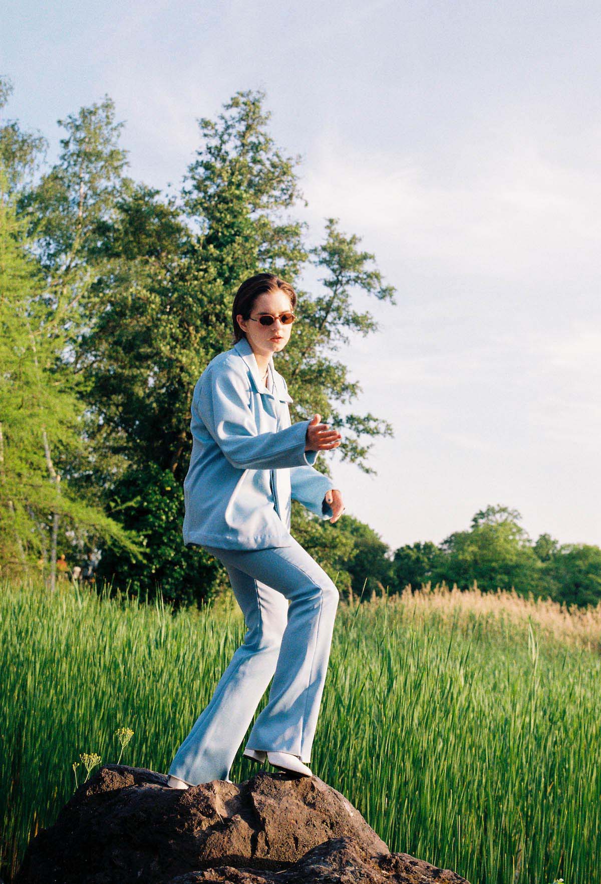 On a rock surrounded by a field of tall green grasses is a young woman in a light blue suit with flared trousers and a wide jacket. Around the field are deciduous trees. Fuffifufzich is wearing white shoes with a wide heel and sunglasses. Her body is slightly turned to the left. She stands on her left leg, the right one is slightly bent and stands higher on the rock. Her right arm is also bent and her hand is open, the forearm points parallel to the ground to the right side of the picture. She is looking towards her right hand. She holds her left arm a little looser beside her body. She wears short brown hair combed back.