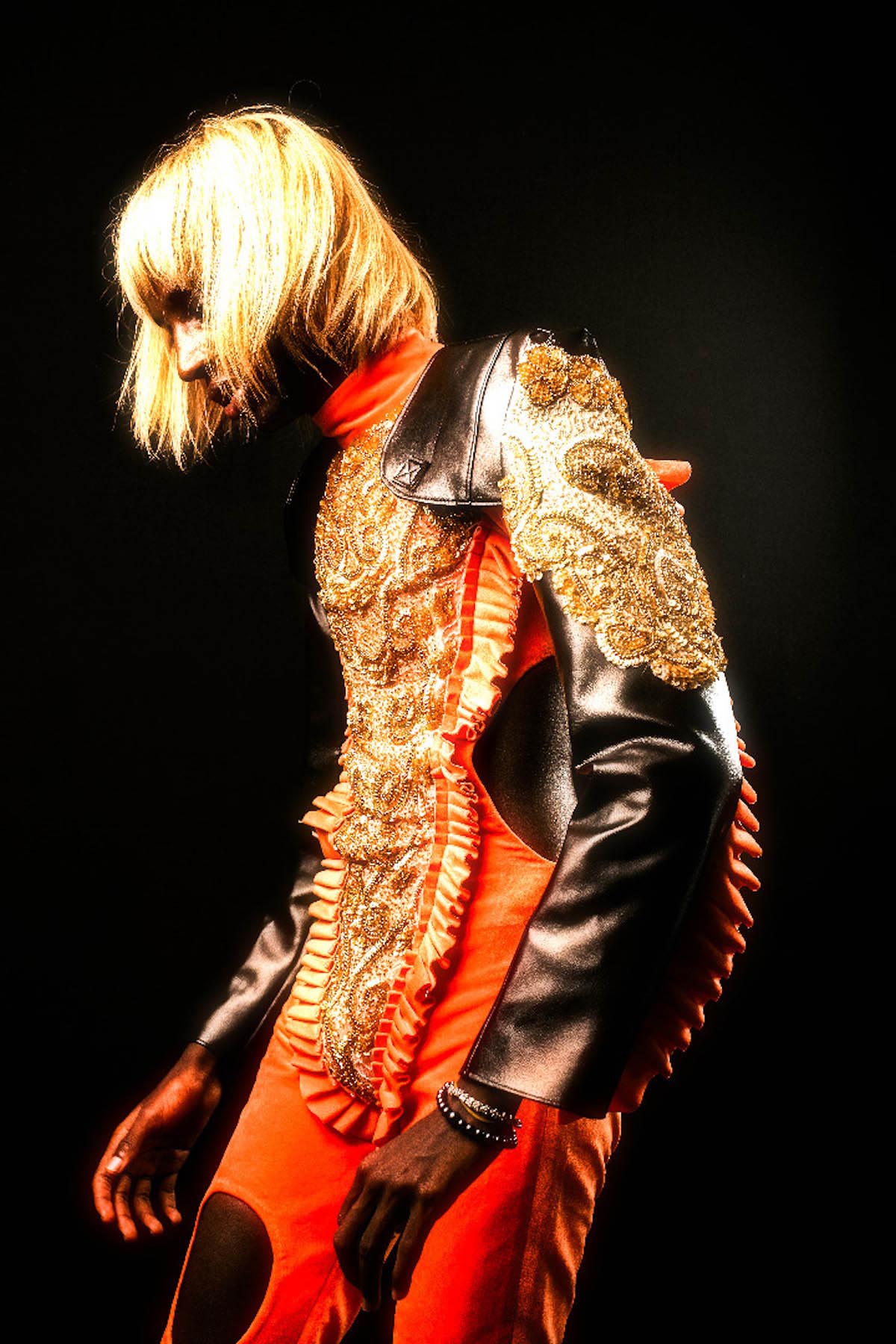Person with blond hair that reaches just below the ear and fringes. Person is visible from the side and down to the knees, the face is mostly covered by hair, wears an orange full-body suit with black sleeves and black big dots at the waist and thighs. On the front of the suit, from the collar to the legs, there is a gold large-scale appliqué with ruffles. The gold is repeated at the shoulder. There is a white and a black pearl bracelet on the left wrist.