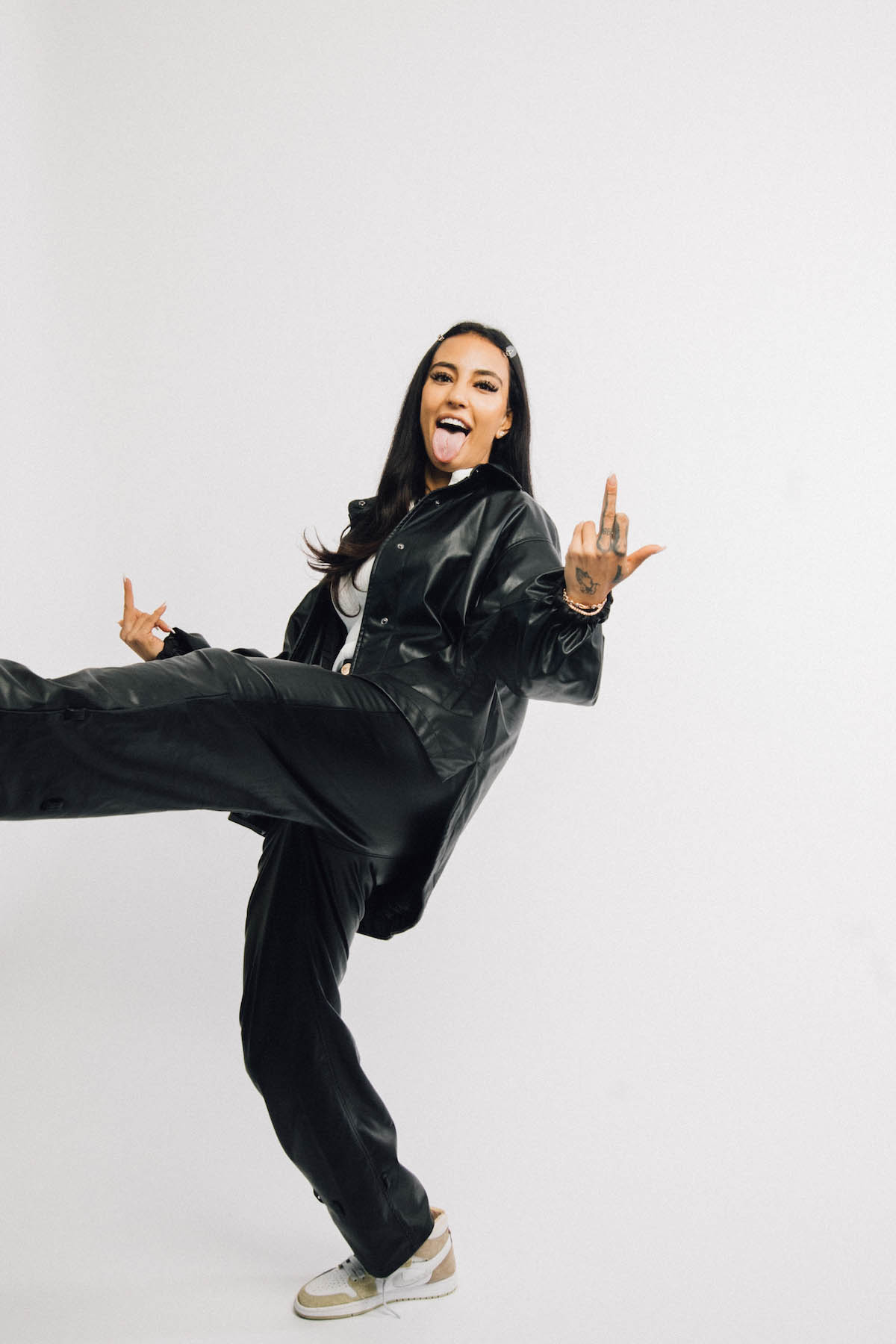 Young woman with long dark hair with a hair clip. She is wearing black oversized trousers and a black oversized jacket with a patent look. She stands on her right leg and swings her left out of the picture. With her left hand she shows her middle finger, on her right she has her index finger and little finger stretched out to form a "mano cornuta". She also sticks out her tongue.
