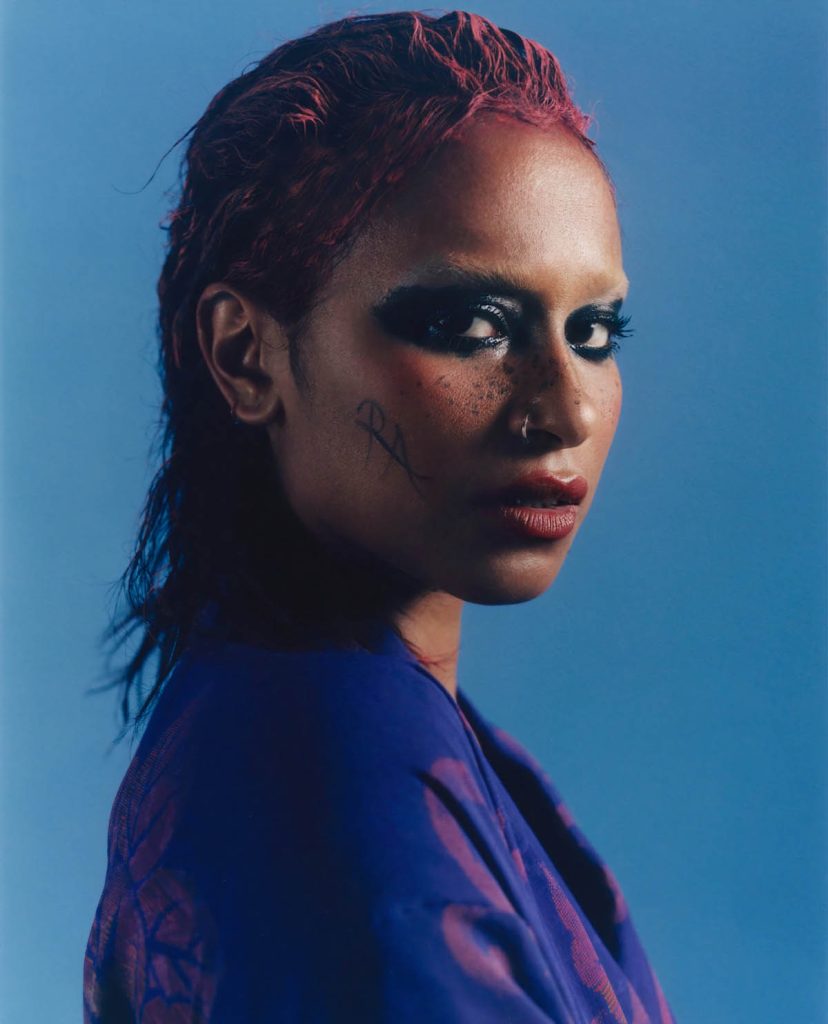 Weibliche Person of Color mit dunkel geschminkten Augen schaut seitlich in die Kamera. Sie trägt einen Nasenring, ein blaues Oberteil mit rote nach hinten gestylten Haare. Auf ihrer Wange ist “RA” zu lesen.