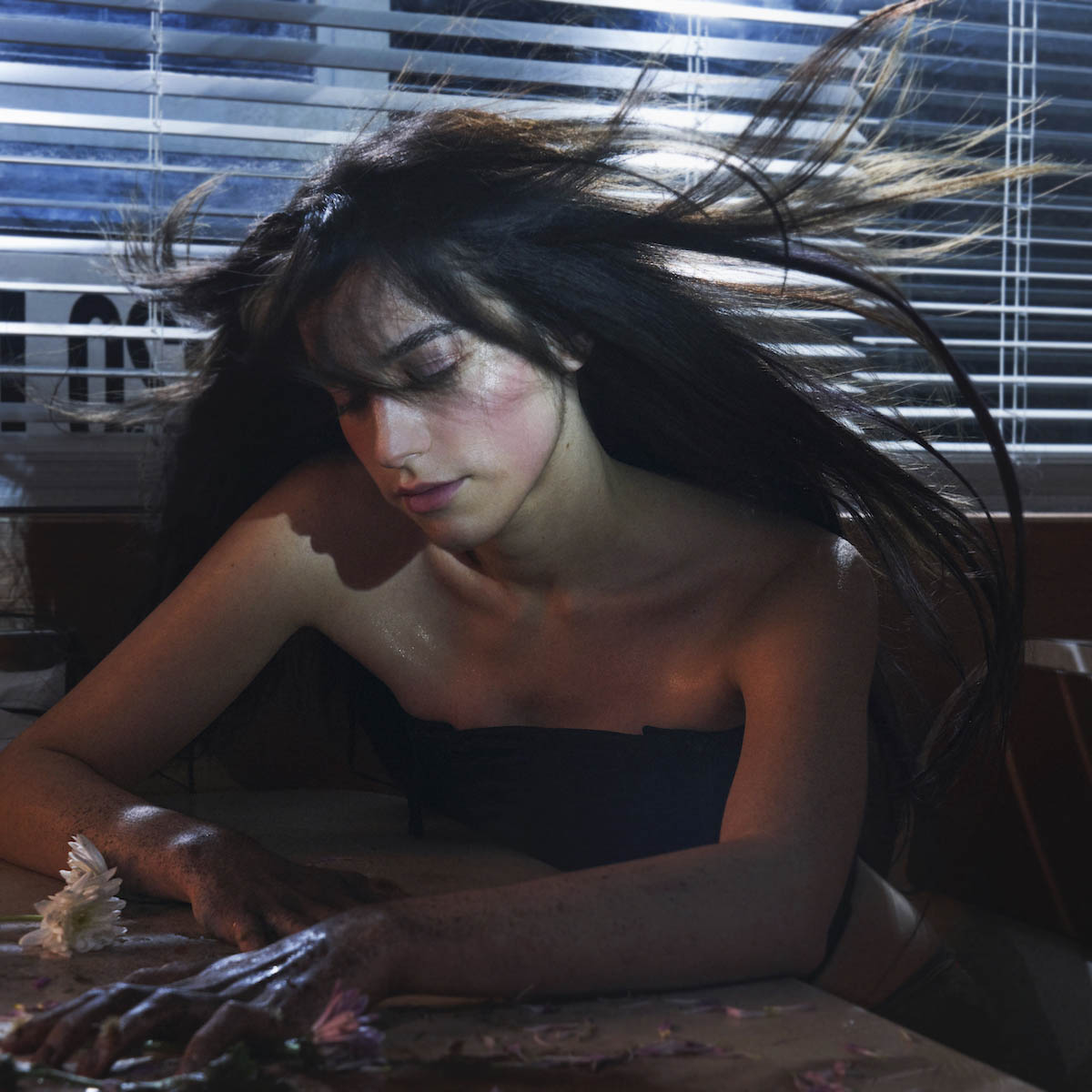 White woman with long brown hair, that's flying in the air, is sitting at a table with her fingertips clawed into the tabletop. She is looking at the table. She is wearing a black top, her shoulders are bare. A half-open blind can be seen behind her. It’s dark outside.