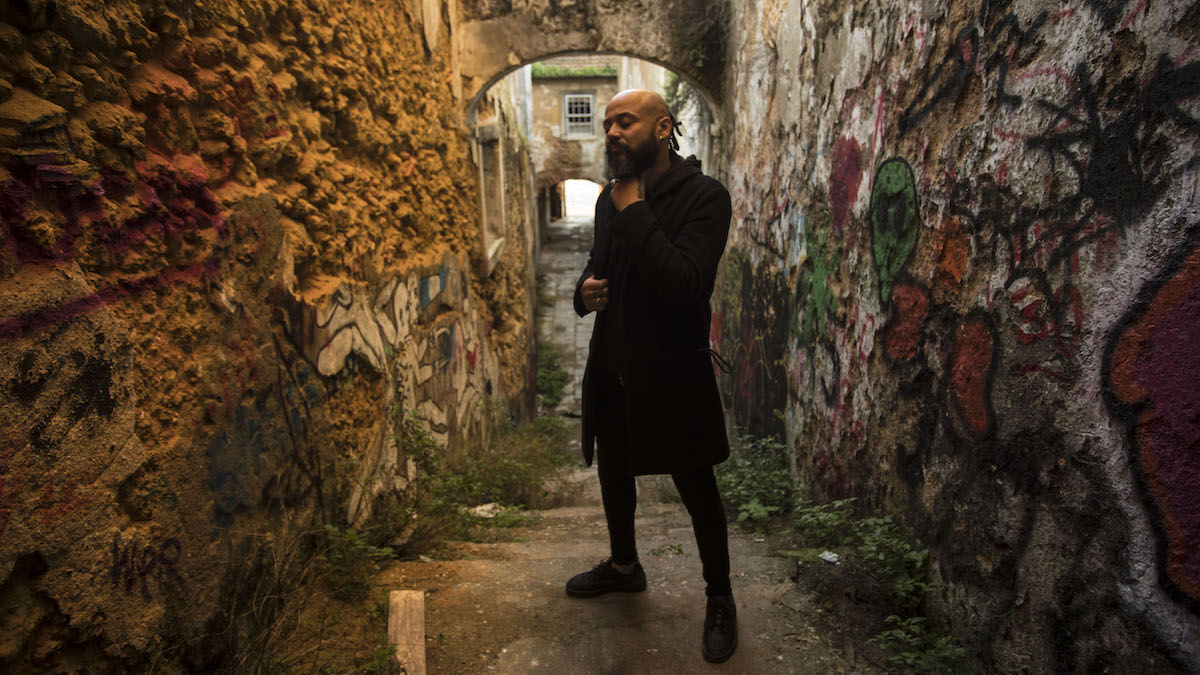 Man with bald head and full black beard in black coat stands between two stone walls sprayed with graffiti. The walls form an alley that leads down steps behind him.