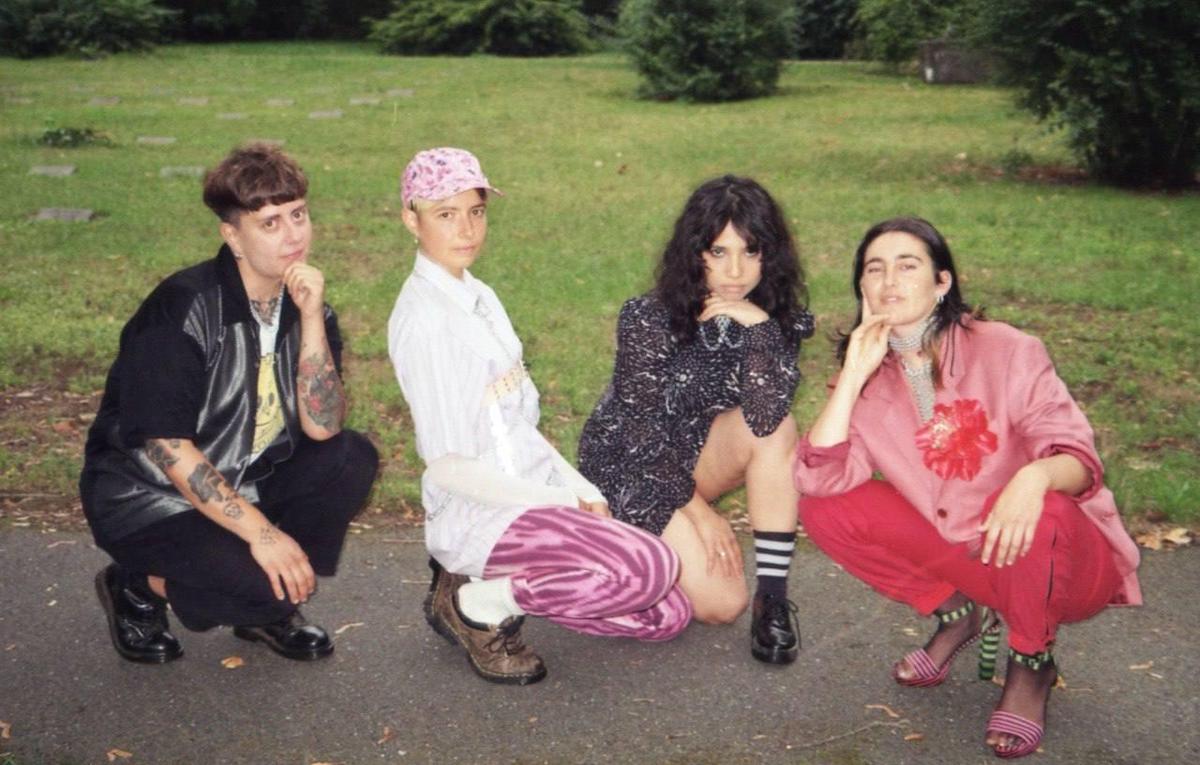 On a paved path in front of a meadow with trees far in the background, four people squat in different positions and look into the camera. The person on the left is wearing a black leather shirt with cloth sleeves over a white T-shirt with a smiley face, black trousers and black flat patent leather shoes. They have short brown hair and tattoos on both arms and rest the left arm on the bent left leg, leaning the closed hand against their chin. In the picture to the right, another person is squatting with a white long blouse, pink and rose leggings, a pink visored cap with pink and purple flowers under which her hair is not visible, white socks and brown flat shoes. The arms rest on the legs. The third person in the picture has long brown curly hair, wears a short black and white dress, black socks with white ringlets and black flat shoes. She rests her head on the back of her left hand and her arm on her knee. The fourth member of the band Taffee has brown straight long hair tucked behind her ears. They are wearing red trousers, a pink blazer with an oversized red flower on the lapel, high heeled shoes with a green and black striped heel and straps. The rest of the sandal is striped pink and black. They spread two fingers of the right hand and leans the hand against their cheek.