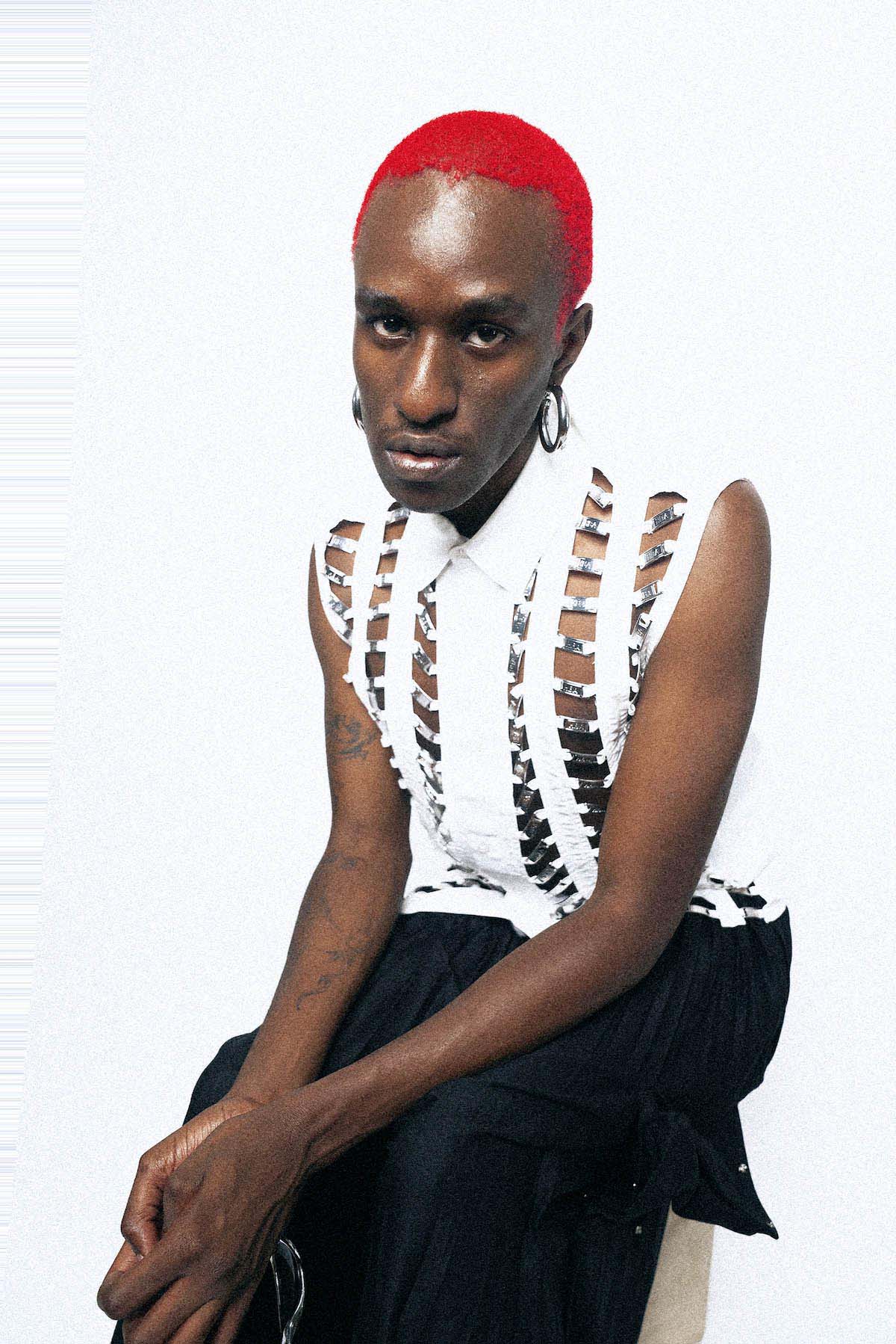 Black person wears black pants, plus sleeveless white blouse, which has silver applications and many cutouts. Tama Gucci wears silver creoles in their ear and has very short red dyed hair. They are sitting, looking directly at the camera and have their forearms resting on their legs with their palms interlocked.
