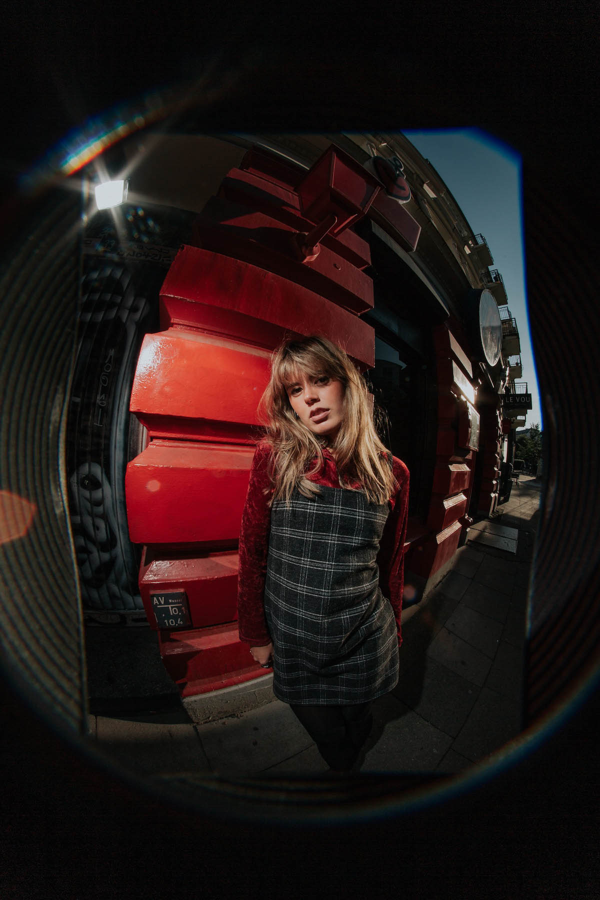 View through a so-called fisheye lens of a white young woman with blond long hair standing in the centre of the picture and looking into the camera. She is wearing a grey and white checked dress with red velvety sleeves. She is standing in front of a red building with balconies and blue sky to the right. The background curves more and more due to the fisheye optics the further it is from the centre of the picture.