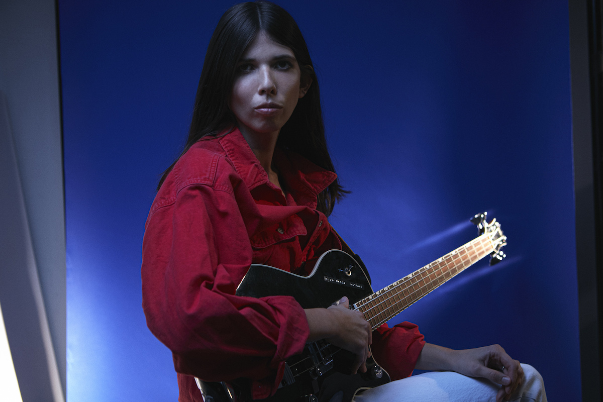 White woman with black hair sits sideways in front of a blue background and turns her head slightly to look at the camera. She is wearing a red denim jacket and white trousers, has her right arm around a black electric bass guitar resting on her leg. Theodora also rests her left hand on her leg.