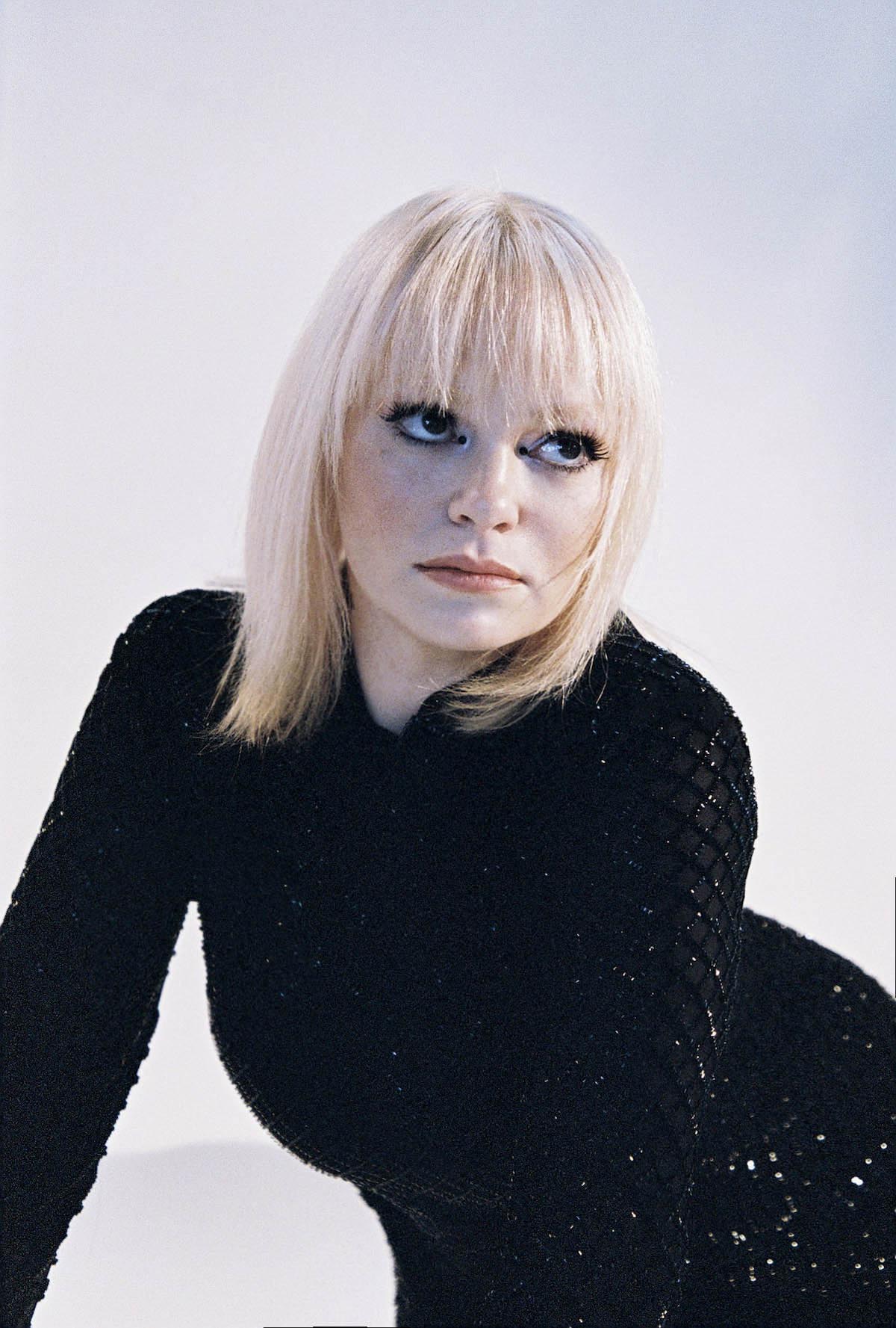 Female person with blond shoulder-length hair and bangs looks to the right side of the picture and up. She is wearing a black sparkly skin-tight dress with long sleeves.