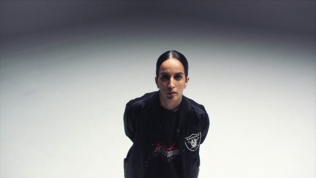 Ebow is wearing a black jacket with a Las Vegas Raiders football team logo patch on the chest and a black T-shirt with red and grey and white lettering underneath. She has her black hair tied tightly back with a centre parting. Ebow is slightly bent forward, has her arms folded behind her back so that they are not visible and is looking at the camera. The background of the picture changes from white to black at the top of the picture.