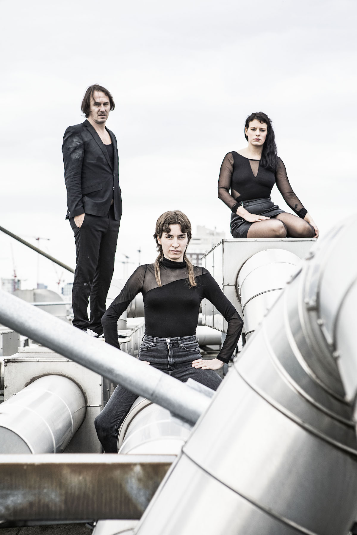 The band Gewalt poses on a flat roof with thick, chrome-coloured ventilation pipes. The sky is grey. Everyone is wearing black clothes, looking sternly into the camera, their faces appear pale. Singer and guitarist Patrick Wanger stands on the left side on a box connecting the pipes and has his hands in the pockets of his suit trousers. He wears a black T-shirt, and a black jacket with white chalk stains on the sleeve. His brown hair reaches below his earlobes. To his right, guitarist Helen Henfling kneels also on a box, in a skin-tight short dress with see-through long sleeves. Her long black hair rests on her left shoulder. Between the two and further front, bassist Jasmin Rilke is sitting wide-legged on a vent pipe. She wears black jeans, a high-necked, long-sleeved top with one shoulder and one arm transparent. Her dark blonde hair is short, only two thick strands are visible at the back of her head, each draped forward over her shoulder. Her hands are each resting on her thighs, her elbows pointing outwards.