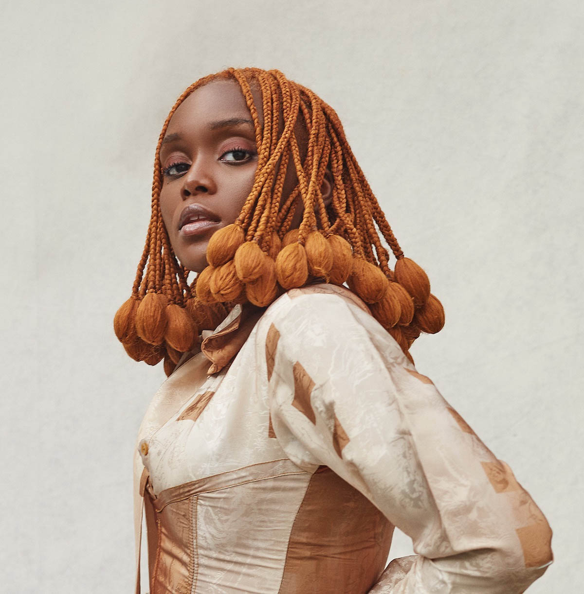 Falana can be seen up to her waist and from the side. However, she turns her face towards the camera. Her hair is orange and braided into many small braids, at the ends of which the hair is formed into a kind of small balloon. She is wearing a cream and brown silk top.