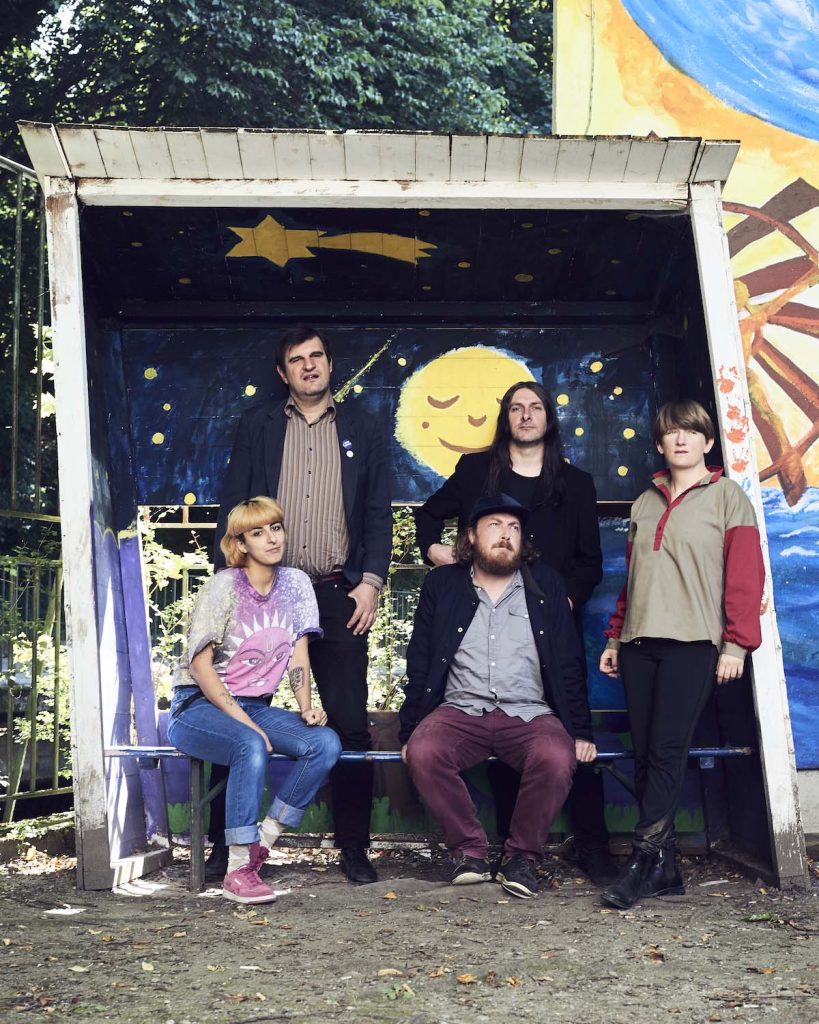 The picture shows the five-member band Friends of Gas. Pictured from left to right are guitarist Veronica Burnuthian, drummer Erol Dizdar, guitarist Thomas Westner, bassist Martin Tagar and singer Nina Walser. Two of the people are sitting and three people are standing in front of a small bus shelter painted white on the outside and made of wood. The interior is blue and painted as a night sky with a yellow shooting star and a moon with a face.