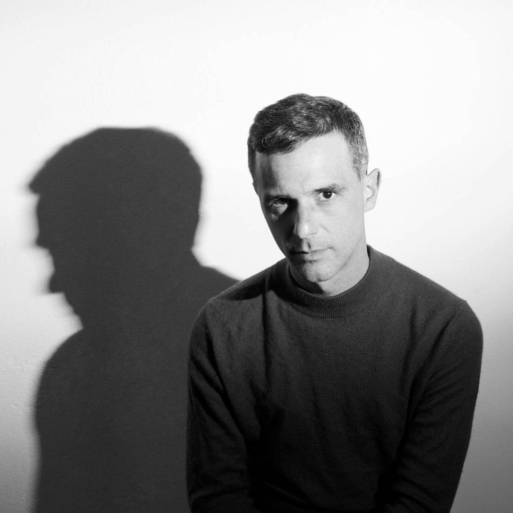 Black-and-white semi-close-up of a white man with short hair looking directly into the camera. Jonas Wehner alias Warm Graves wears a monochrome sweater, to the left of him his silhouette can be seen as a shadow.