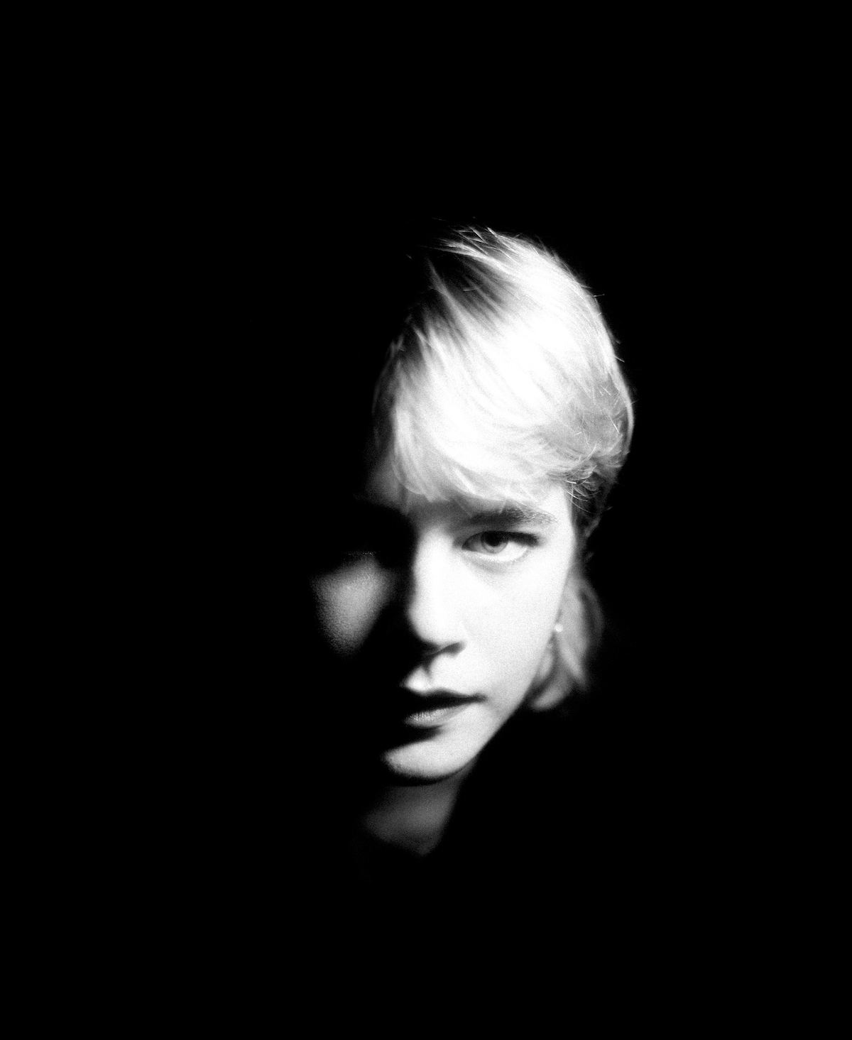 On a black-and-white picture, with a deep black background, the right half of Zaho de Sagazan's face is the most visible. A black shadow only hints at the left half. Her blond hair falls loosely into her forehead. She looks into the camera.