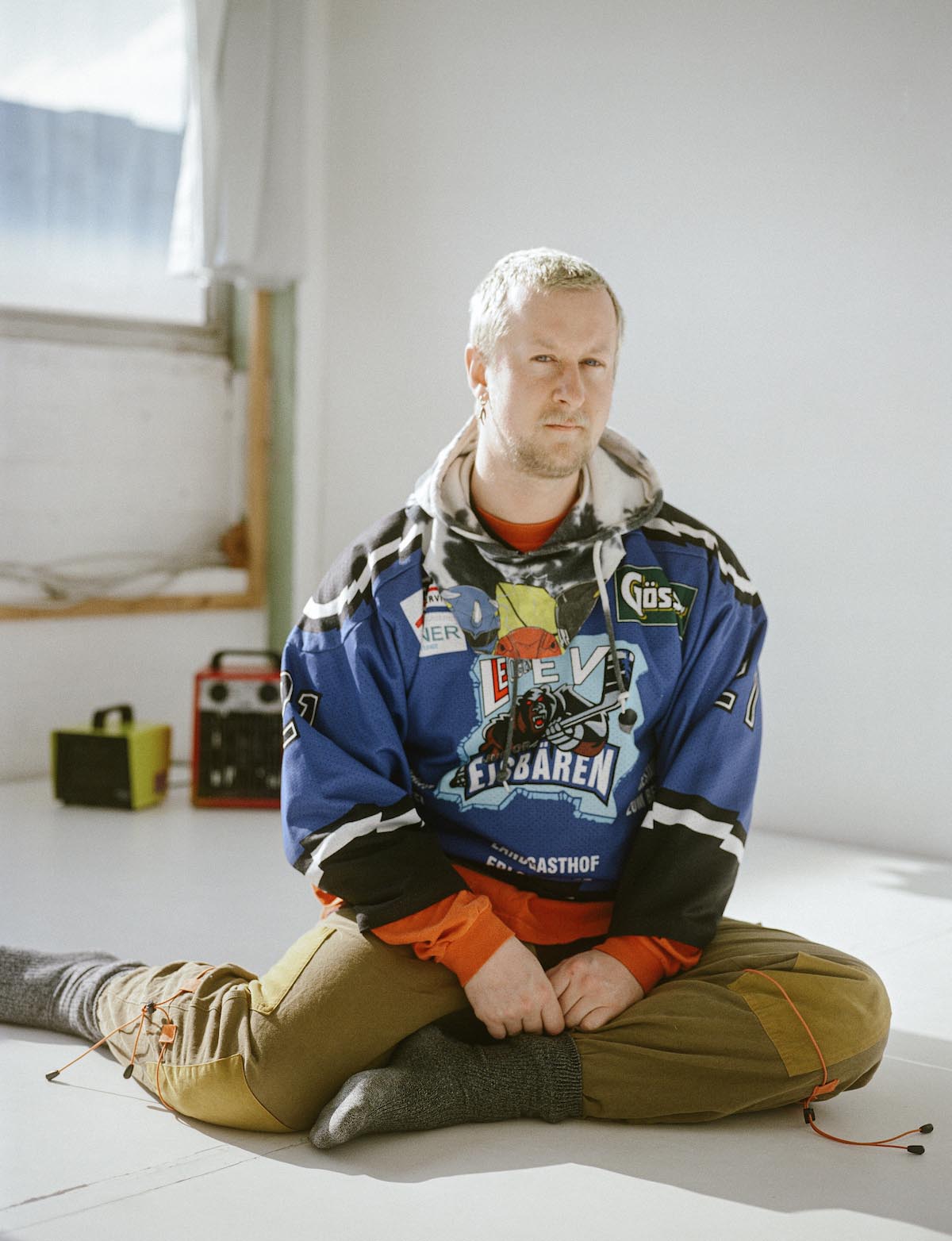 Long shot of Colin Self. Self is sitting upright on the floor in a half cross-legged position, but the right leg is positioned outwards and slightly bent. Self's hands rest on the calf of the left leg, which is bent inwards. Colin Self is wearing a black and white batik hoodie, over it a blue, black and white jersey of an ice hockey team with the name "Polar Bears", brown long trousers with orange thin bands around the knees and grey, thick woollen socks. Self has short blond hair, a blond three-day beard, a long gold earring in his right earlobe. Colin Self is sitting in a room with white walls, windows to the left.