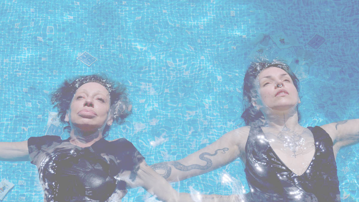 Two white women with brown hair lie flat next to each other in a light blue tiled pool, floating in the water, only their faces are not covered with water. They look up, both wearing black clothes. They are Little Annie on the left and Evilyn Frantic on the right. Light is reflected in the water. They can both be seen up to their waists and have their arms spread out to their respective sides. Evilyn has a snake tattooed on the inside of her right arm and wears a septum piercing.