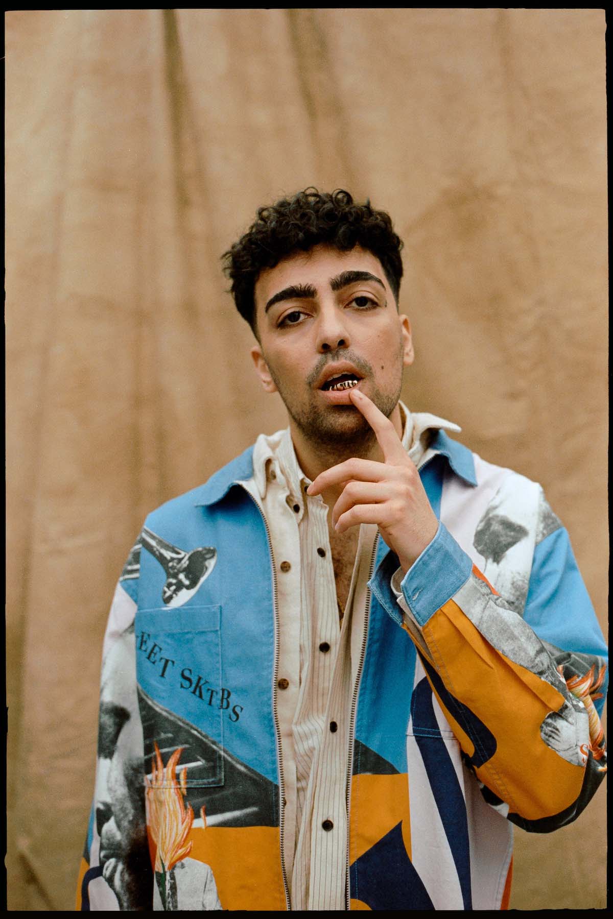 The rapper BRKN can be seen up to his waist against a brown fabric background. He has black, short curly hair, dark eyes, prominent black eyebrows, a three-day beard and he wears a blue-orange-white jacket with pictures of a torch, trumpet and a trumpet player. He looks into the camera, has his mouth slightly open and puts his left index finger to his lower lip. You can see a golden grill on his lower teeth.