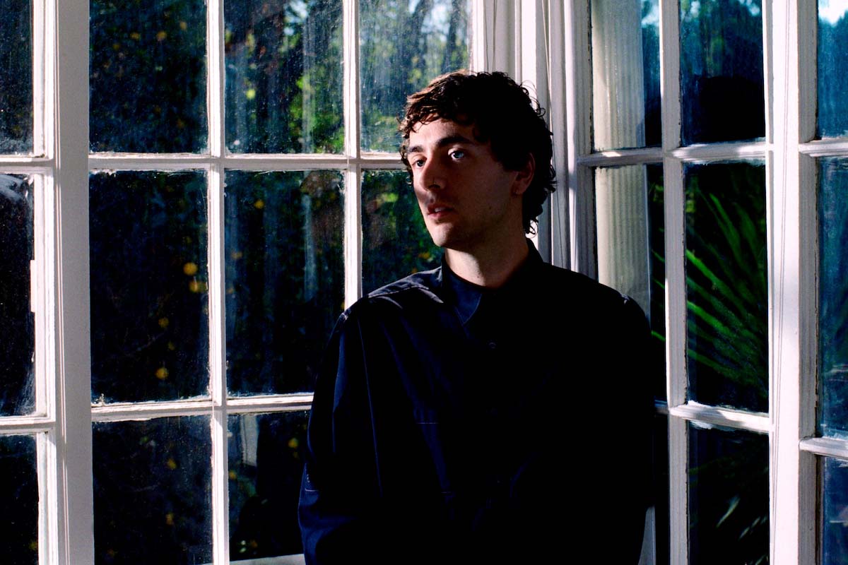 White, young man with curly short hair can be seen up to the waist, Casey MQ is wearing a dark blue, long-sleeved, buttoned-up shirt. His mouth is slightly open, he is looking out of the picture to the left, he has a nose ring. He is standing in the corner of the window front of a house. The panes are divided by many bars. Light comes from outside and casts shadows on his face. In the background, a lemon tree and other Mediterranean plants can be glimpsed.