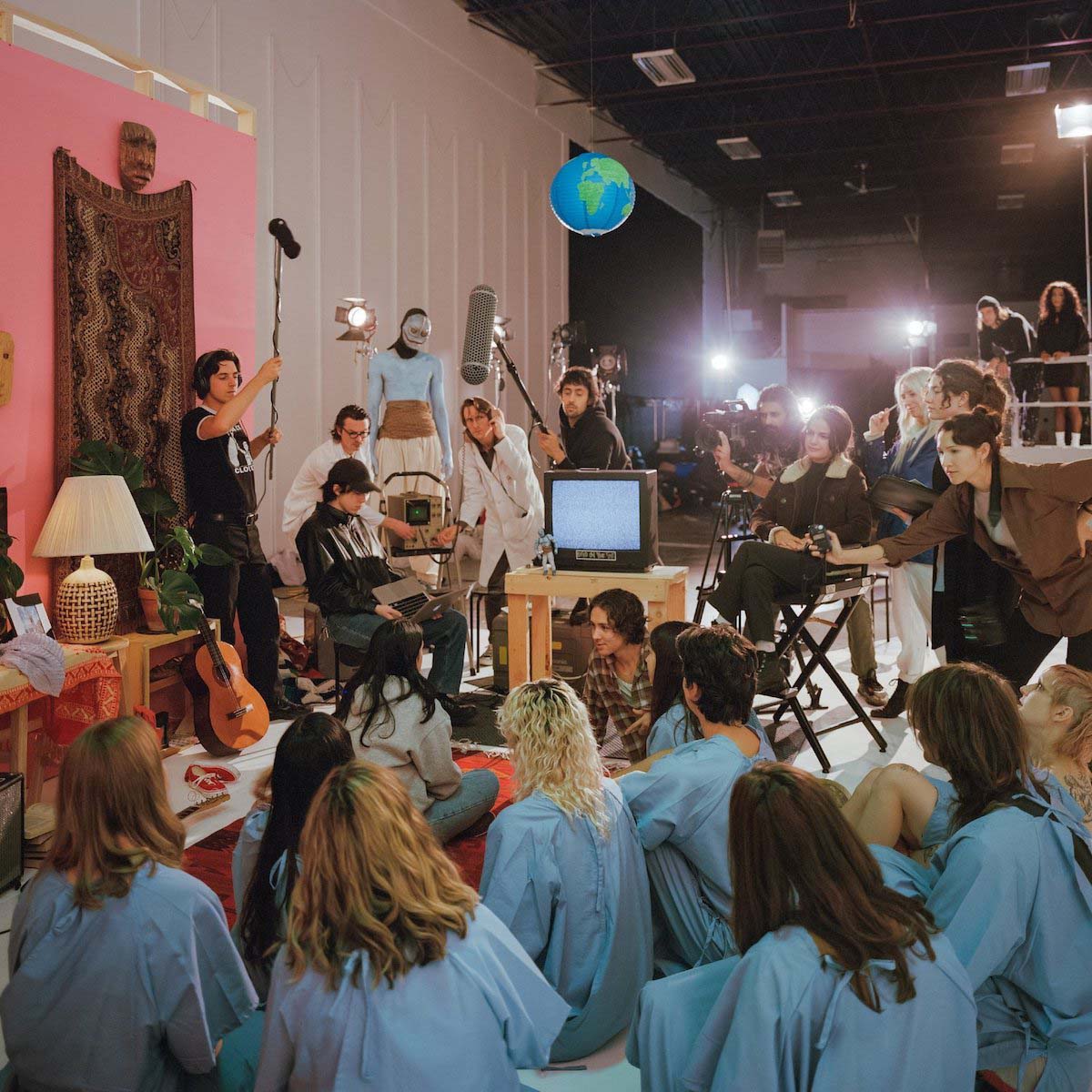 Scene with numerous people in a youth room with lamp, tapestry, TV, guitars that has been set up in a corner of a large film studio. The aesthetics of the image can also be found in the Crack Cloud video for "Please Yourself". In the foreground, a group of people in blue patient gowns sit on the floor. They can all be seen from behind watching two people in everyday clothes, also sitting on the floor talking, with a camera pointed at them. In the background is a film crew consisting of, among others, the director on the director's chair, people for camera, sound and lighting. As well as two other people in white coats and one with a monster mask.