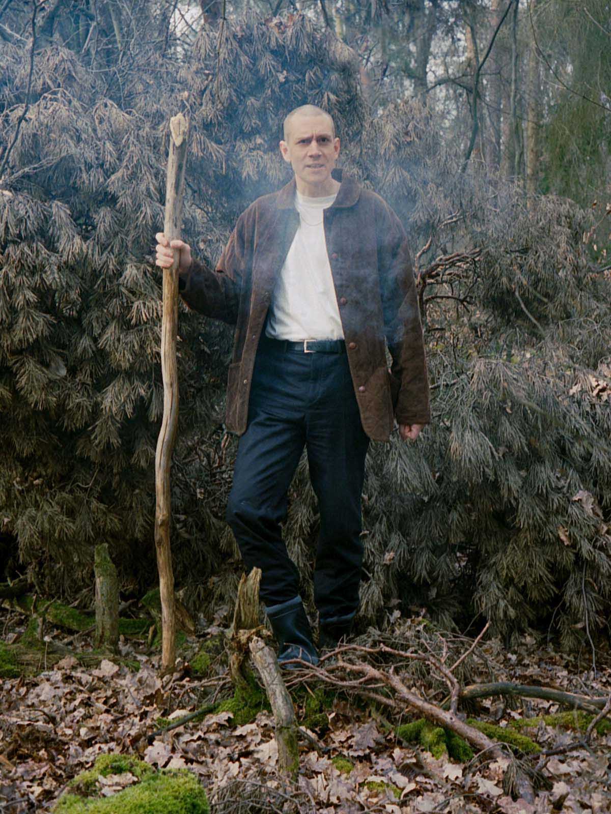 White, middle-aged man with shaved bald head, white T-shirt, dark blue jeans and brown jacket leans on a branch in his right hand as if on a walking stick. Das Kinn is in the forest in front of a bushy branch with many twigs and needles.