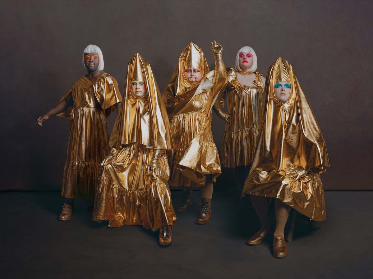 Five people in different golden, knee to ankle length dresses and golden shoes, standing side by side and posing towards the camera in a dynamic and exalted manner. The person on the far left and the second from the right are wearing white bob wigs, the other three have on golden cone-shaped headdresses tapering well above the head with expansive fabric that hugs the face and falls over their torso. All five members of Drag Syndrome wear eye-catching make-up, especially the eye shadow, which is a different colour on each of them.