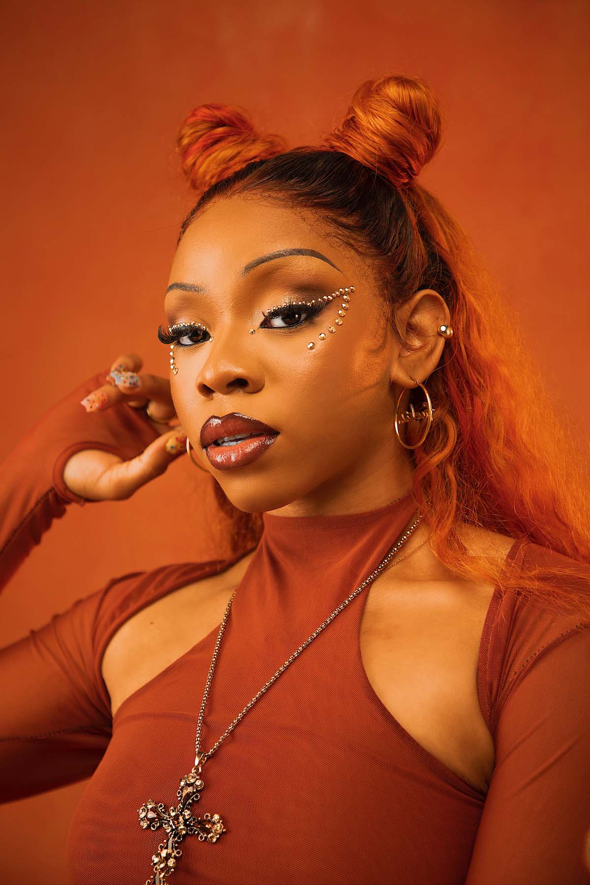 Close-up image in monochrome colours of a young Black woman looking at the camera, against a reddish brown background. She wears a long-sleeved, tight top with cutouts on the collarbone in the same colour. Fave's red hair is styled into so-called double buns at the top of her head, with the rest of her long hair falling down her back. She wears glitter stones in the shape of cat's eyes around both eyes, gold creoles and a silver chain with an intricately designed large cross. Her lips are outlined with brown lipliner and glossed. Fave poses with her right hand raised to her right ear, her long, colourfully painted fingernails standing out.