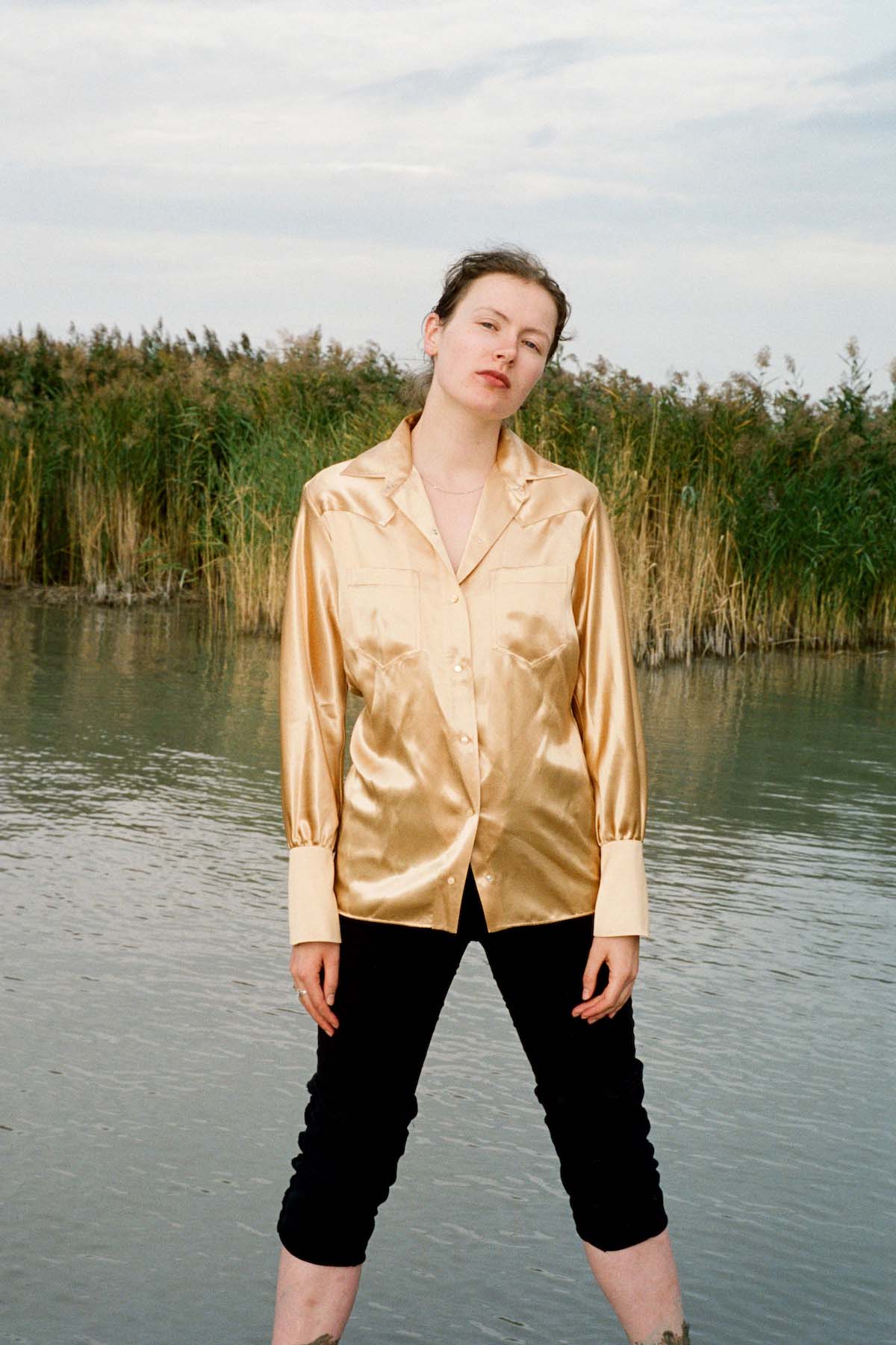 Young, white woman stands with wide legs in or in front of the edge of a lake, her feet are not visible. Water stretches in the background, closed off by green-brown reeds. Rosa Anschütz has tied her brown hair into a plait, wears a loose, golden blouse and black shin-length, tight trousers. She lets her arms hang close to her body and looks confidently into the camera.