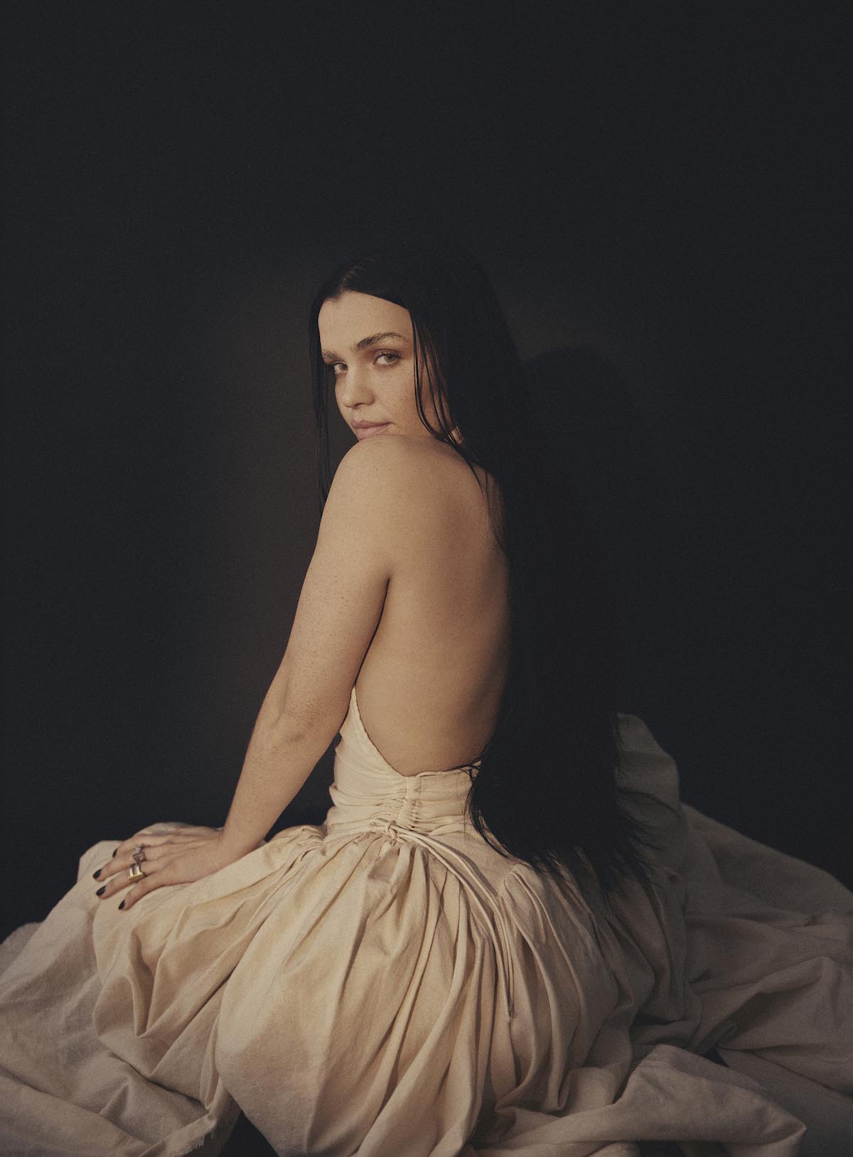 Young, white woman sitting on the floor is seen from behind and turns her head to look at the camera over her left, naked shoulder. ARY has long black hair that blends into the black background. She wears a cream-coloured, backless, shoulderless dress draped over her legs and the floor. She rests her left hand on her left leg.