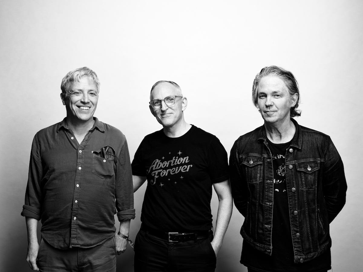 Black and white photo of the band Codeine, three older, white men standing next to each other and smiling at the camera, only the man on the left shows teeth. He wears short, grey hair, a denim shirt with reading glasses peeking out of the chest pocket, he sticks his thumbs in his trouser pocket. In the centre is a man wearing glasses and a black T-shirt that says "Abortion Fever". He has one hand in his pocket. The third man has combed back neck-length hair, is wearing a T-shirt, an open denim jacket and has both hands behind his back.