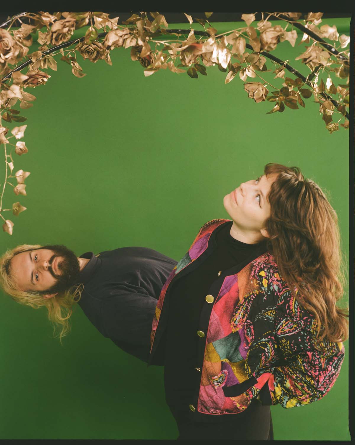 The band Freelove can be seen against a green background, above the woman and the man are branches with golden leaves and flowers. The woman stands on the right, wears a colourful blouson with golden buttons over her otherwise black clothes, has her hands in her trouser pockets and looks up at the branches. Her long brown hair falls down her arms and back. The man stands behind her and looks into the camera, only his upper body can be seen, which he bends almost horizontally to the left. He has a full black beard and a blond-dyed mullet.
