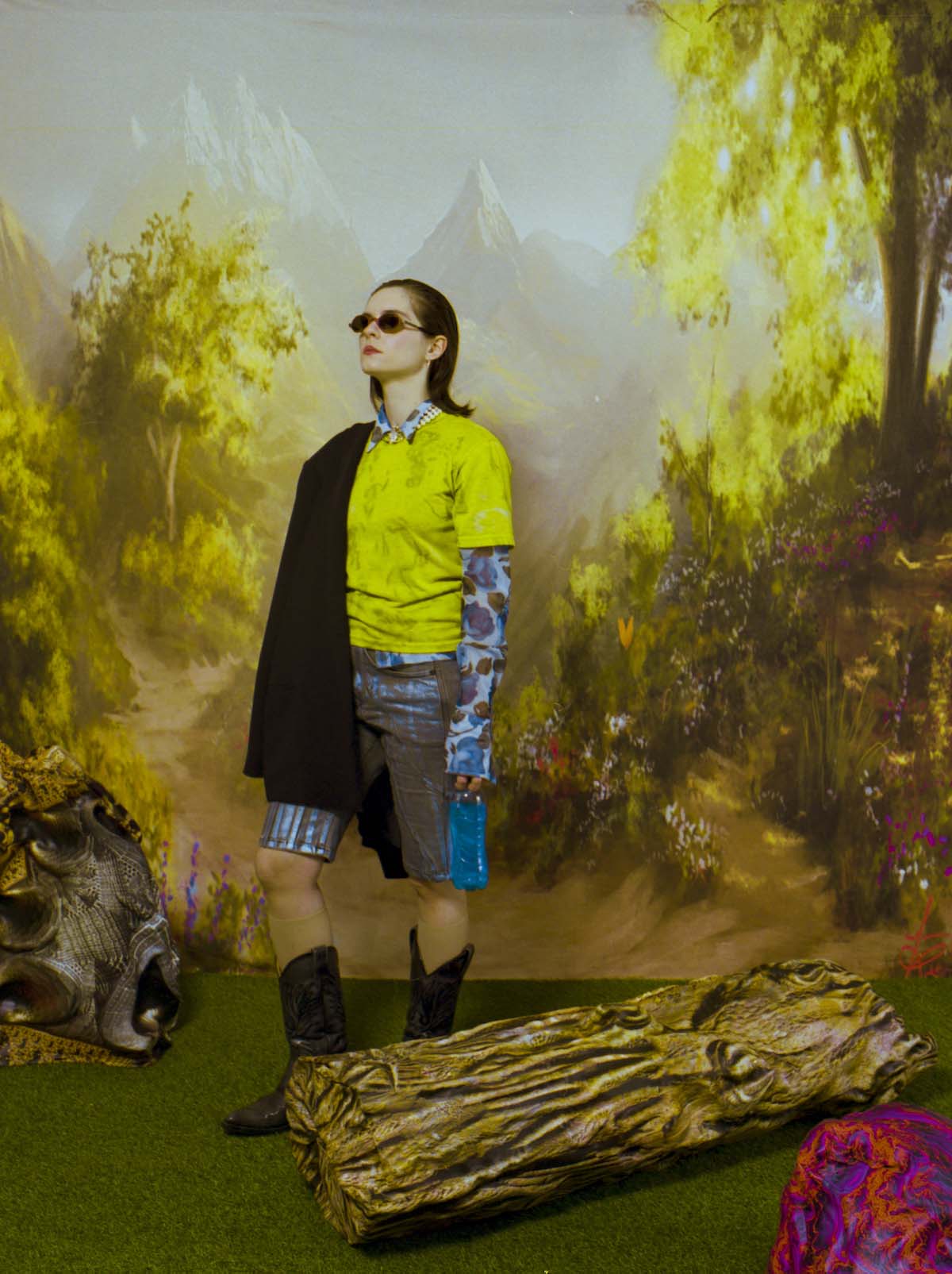 Fuffifufzich stands upright with cowboy boots, short blue-brown trousers, a long-sleeved shirt in the same colours over which she wears a yellow T-shirt, and a black coverlet over her right shoulder in a backdrop in which, among other things, a prop tree trunk lies. The background is a painting showing a landscape with snow-covered mountains, trees, a path and grasses. She is also wearing sunglasses, has her shoulder-length brown hair slicked back and is looking out of the picture to the left.
