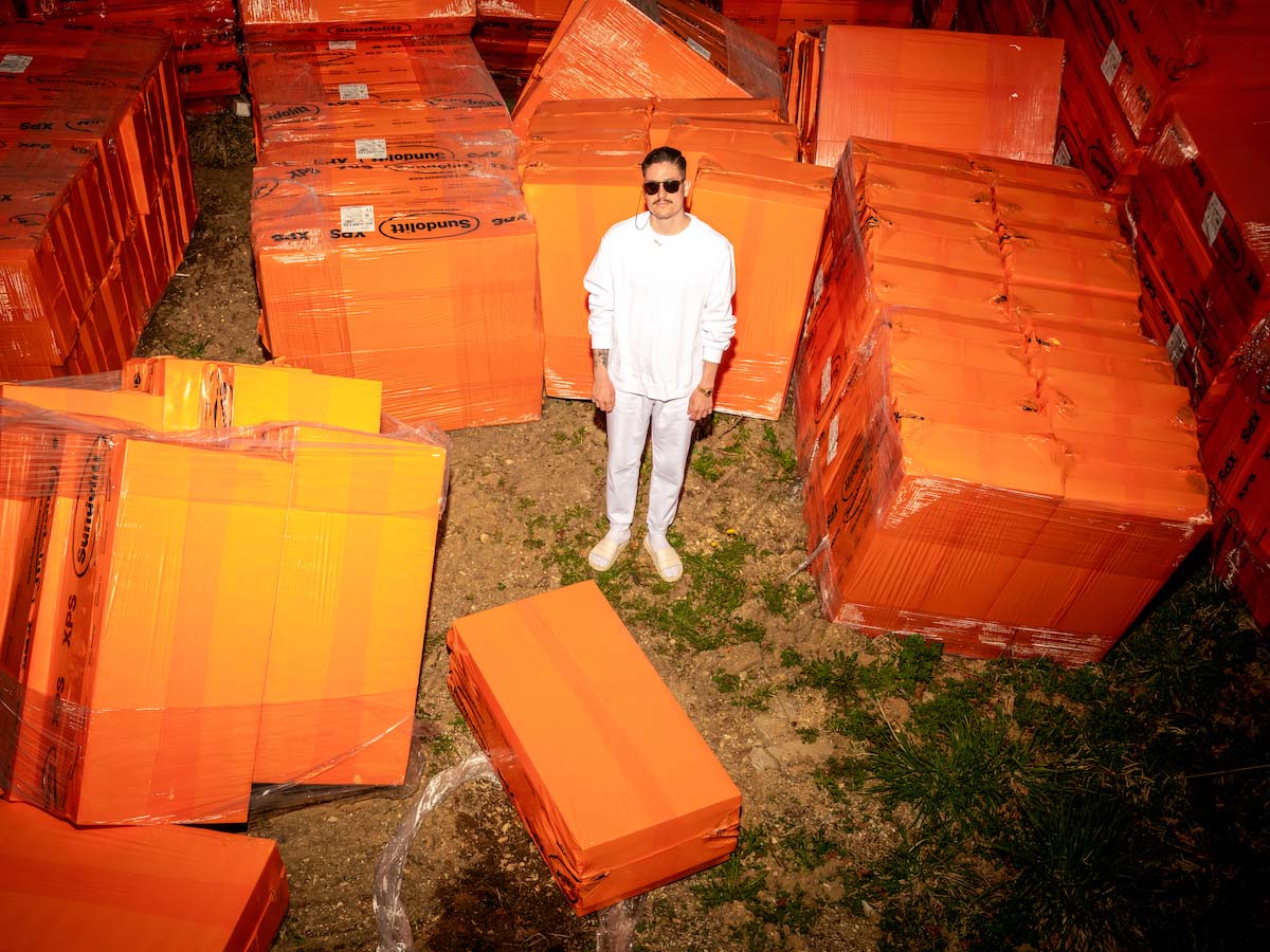 Long shot of Garagen Uwe at a storage place for insulation material. He looks towards the camera and is wearing a white jumper, white trousers, white socks, white slippers, sunglasses and short black hair. He is standing between blocks of material shrink-wrapped in orange foil, which are spread somewhat untidily around him.