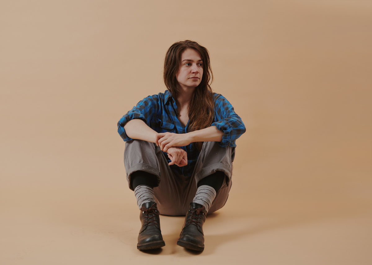 White woman with long brown hair wearing a blue and black plaid shirt, grey trousers with legs folded over, black shoes and grey gauntlets sits on the floor against a light brown background. Nichtseattle's legs are bent, she rests her elbows on her knees and holds her right wrist with her left hand. She looks out of the picture to the right.