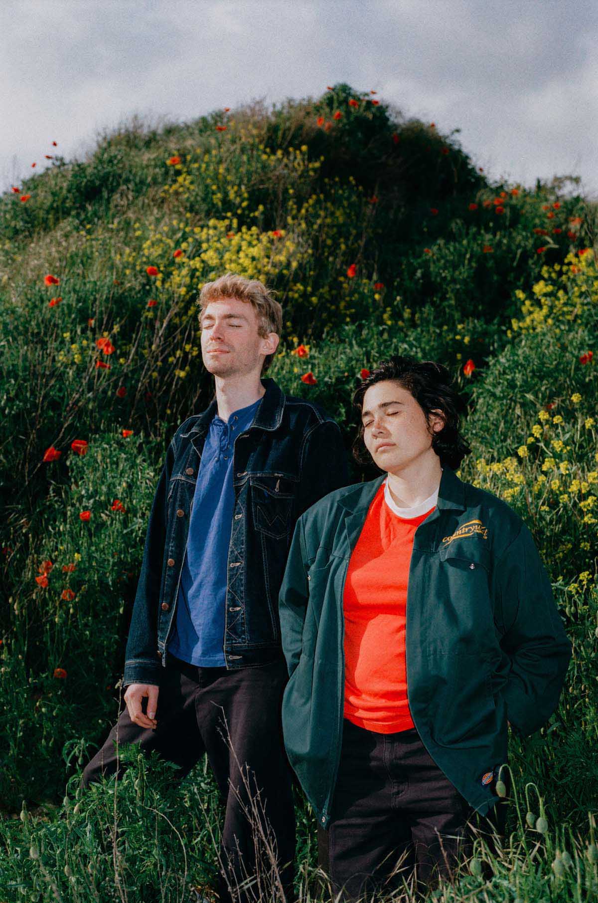 The two members of the band Aili are standing in front of a hill covered with tall grass and flowers with yellow and red blossoms. Aili Maruyama and Orson Wouters are standing upright next to each other in the middle of the picture, their eyes closed and their bodies slightly turned in to the left. He has short blond hair, a three-day beard, wears black trousers, a blue T-shirt and a darker denim jacket. She has chin-length black hair tucked behind her ears on the visible side. Aili is wearing an orange-red T-shirt with a dark green hip-length jacket over it and black trousers.