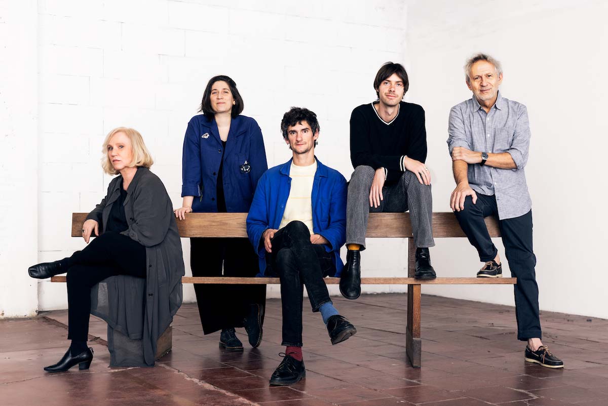 Five, white people look into the camera. The band Aksak Maboul is spread out on and around a bench with a backrest. The blond woman on the left sits on the bench, her legs crossed. To her right, behind the bench, is a black-haired woman, next to whom a man is sitting on the bench again. Among other things, he is wearing a blue shirt and two different coloured socks (red and blue). Next to him, a man sits on the back of the bench and puts his feet on the seat. At the left end of the bench, the oldest of the three men is also sitting on the backrest, with one leg on the floor and the other also resting on the seat.
