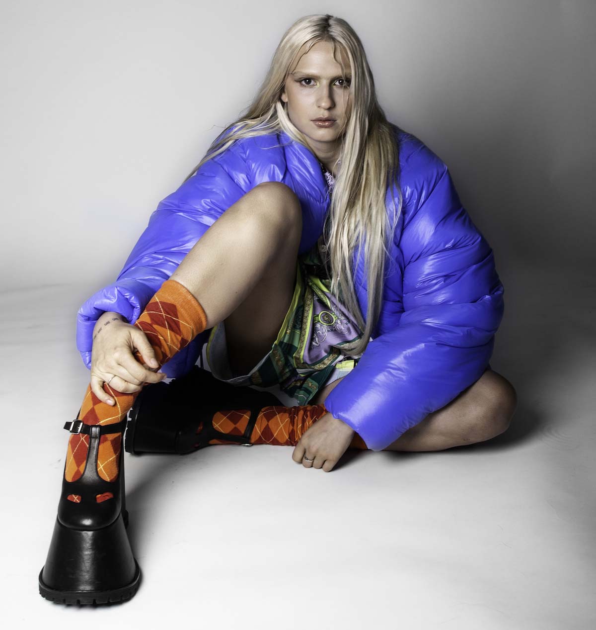 Young, white person with long blond hair sits on the floor in front of an off-white background. Banoffee looks blankly at the camera, has her right leg, which she clasps above the ankle with her right hand, propped up, the other leg is bent horizontally. She is wearing an oversized bright blue quilted jacket, green-purple-yellow patterned shorts, orange-red checked socks and black platform sandals with extremely high and wide soles.