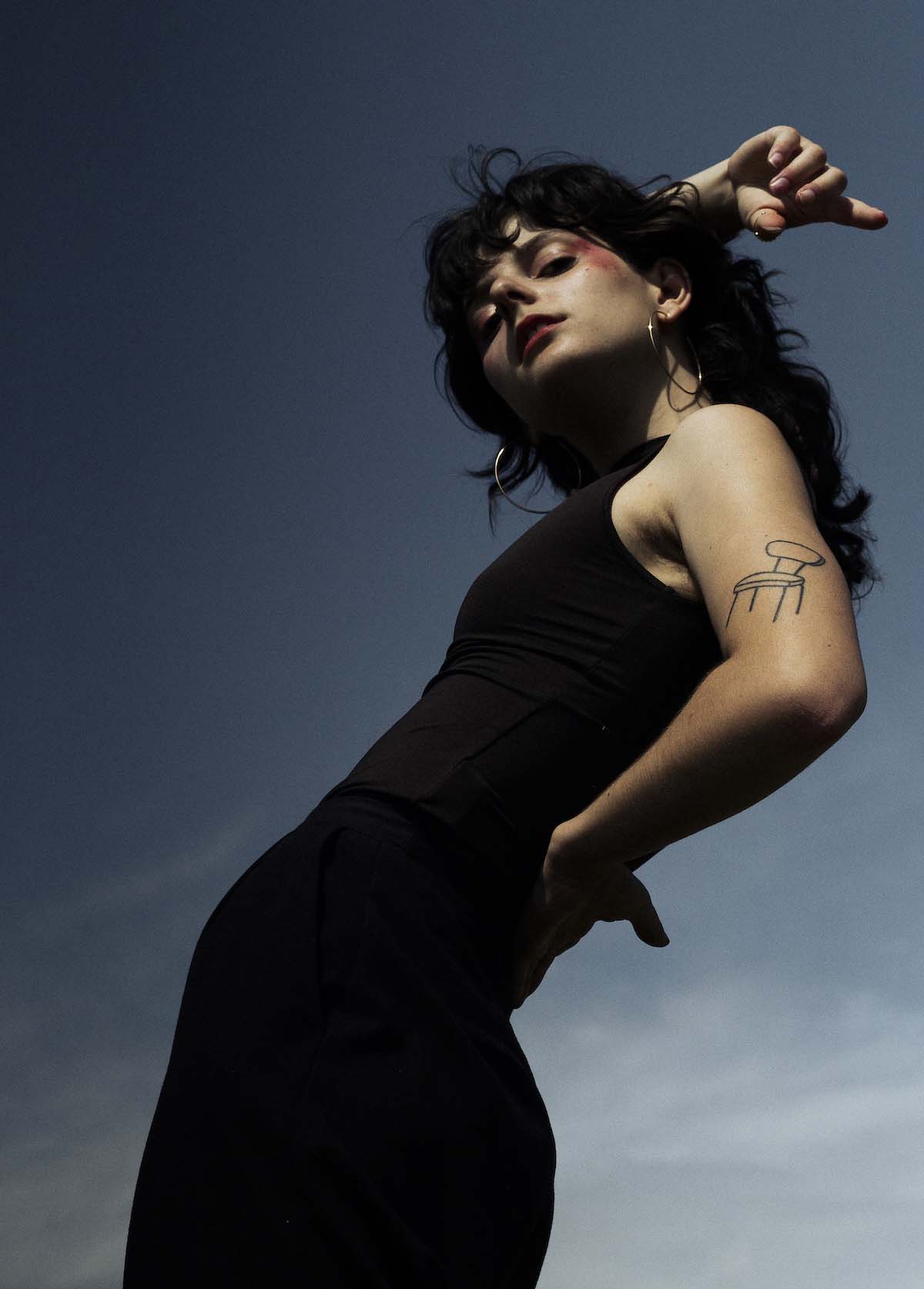 Young, white woman with long, wavy black hair in a black, tight, sleeveless dress is seen from the side and stands leaning back in a model pose. She is looking into the camera, which is photographing her from below. In the background, blue sky with cirrus clouds. C'est Karma poses with her right arm casually above her head, her left hand leaning against her back, thus bringing her upper arm into focus, on which she has a simple chair tattooed.