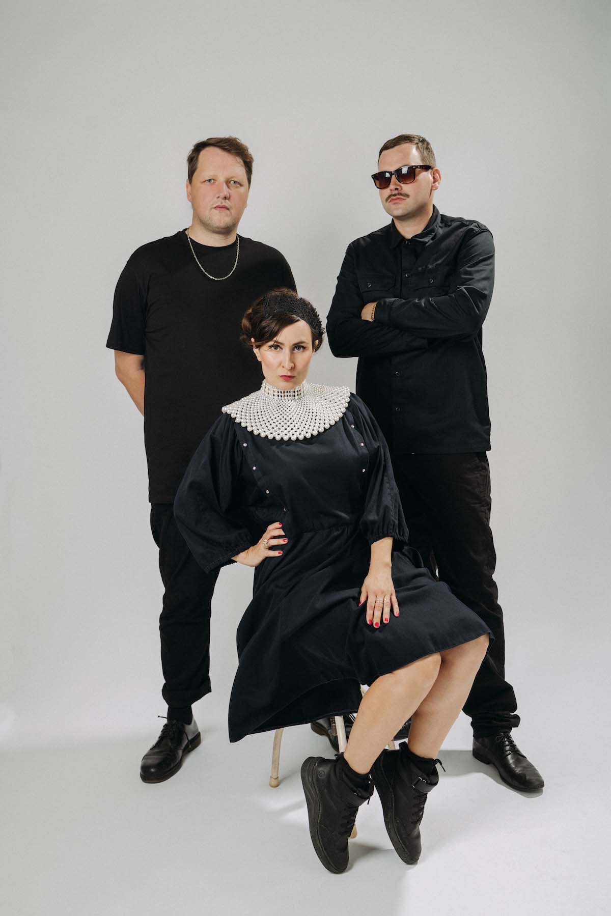 A woman and two men pose for the camera in front of an off-white background. The band Dina Summer is dressed completely in black. The woman in the middle wears a white, flared collar over her black dress and her brown hair in an updo. She sits on a stool in front of the other two, has her right hand resting in her side and the other on her legs that are bent to the side, and she puts her feet in black trainers up on her toes. The one at the back right with short hair has his arms crossed and is wearing a black shirt, sunglasses and moustache. The other one, also with short brown hair, has a black T-shirt on and his arms behind his back.
