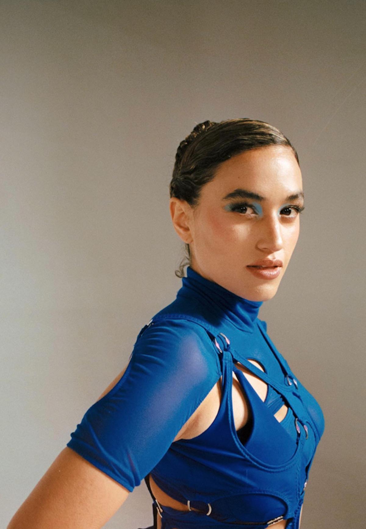 Close-up of Empress Of against a grey background. She is seen from the side and turns her head towards the camera. She is wearing a nifty blue top with a turtleneck, short sleeves and cutouts, in which multi-layered pieces of fabric are held together with rings. She wears matching blue eye shadow. Her brown hair is braided and fixed at the back of her head, her lips slightly parted.