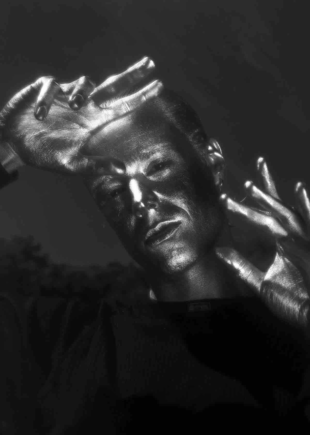 Monochromes Schwarz-Weiß-Porträtfoto von J. Vague, auf dem seine Hände, Gesicht und Hals ganz mit silberner Farbe bedeckt sind. Der Hintergrund ist dunkelgrau. Er hält den Kopf leicht schräg und die Hände filigran vor der Stirn und neben seinem Gesicht. Er trägt ein schwarzes Oberteil.